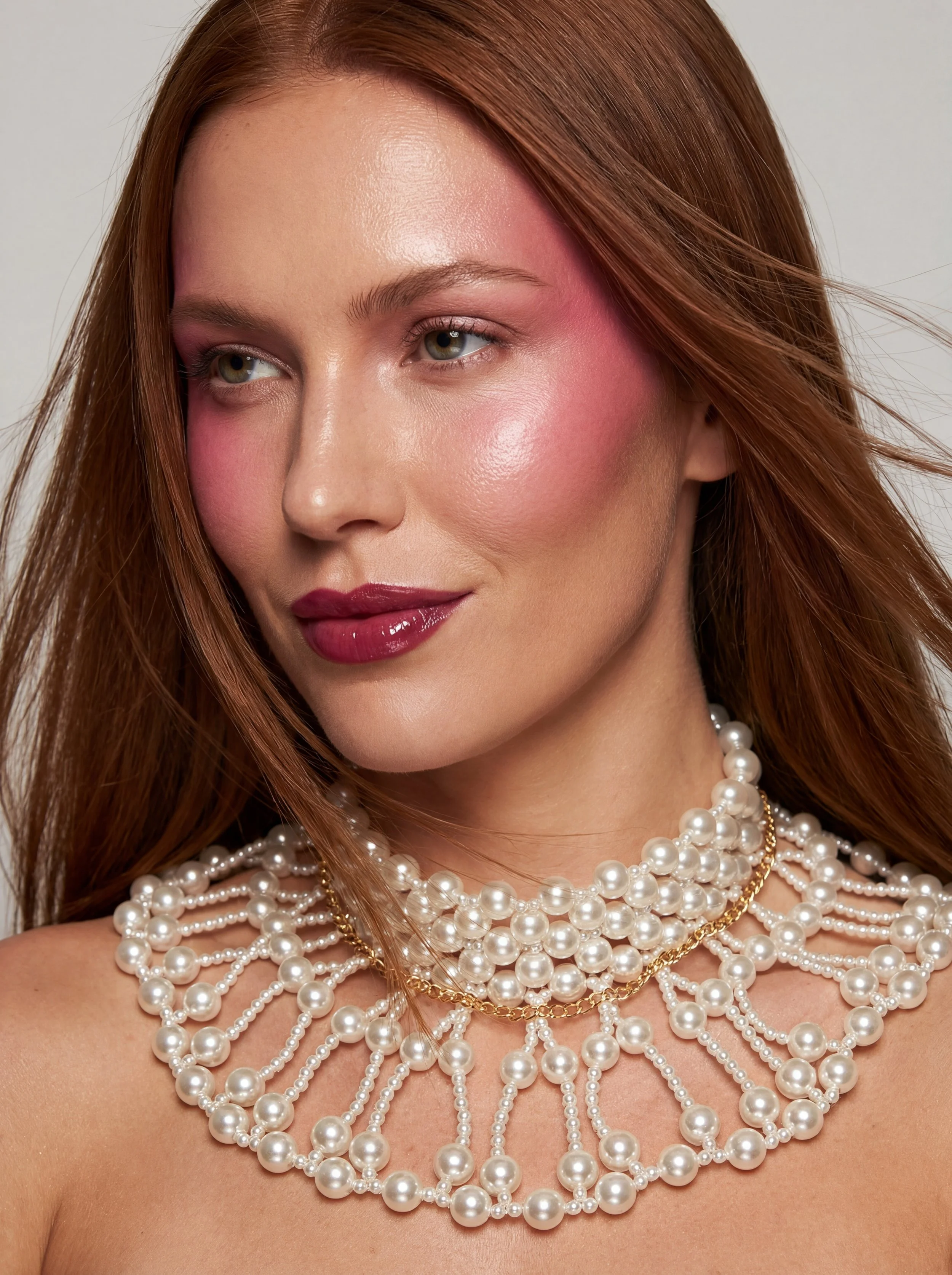 Close-up of a woman with red hair wearing multiple strands of pearl necklaces and a gold chain, with pink makeup on her cheeks, lips, and around her eyes.