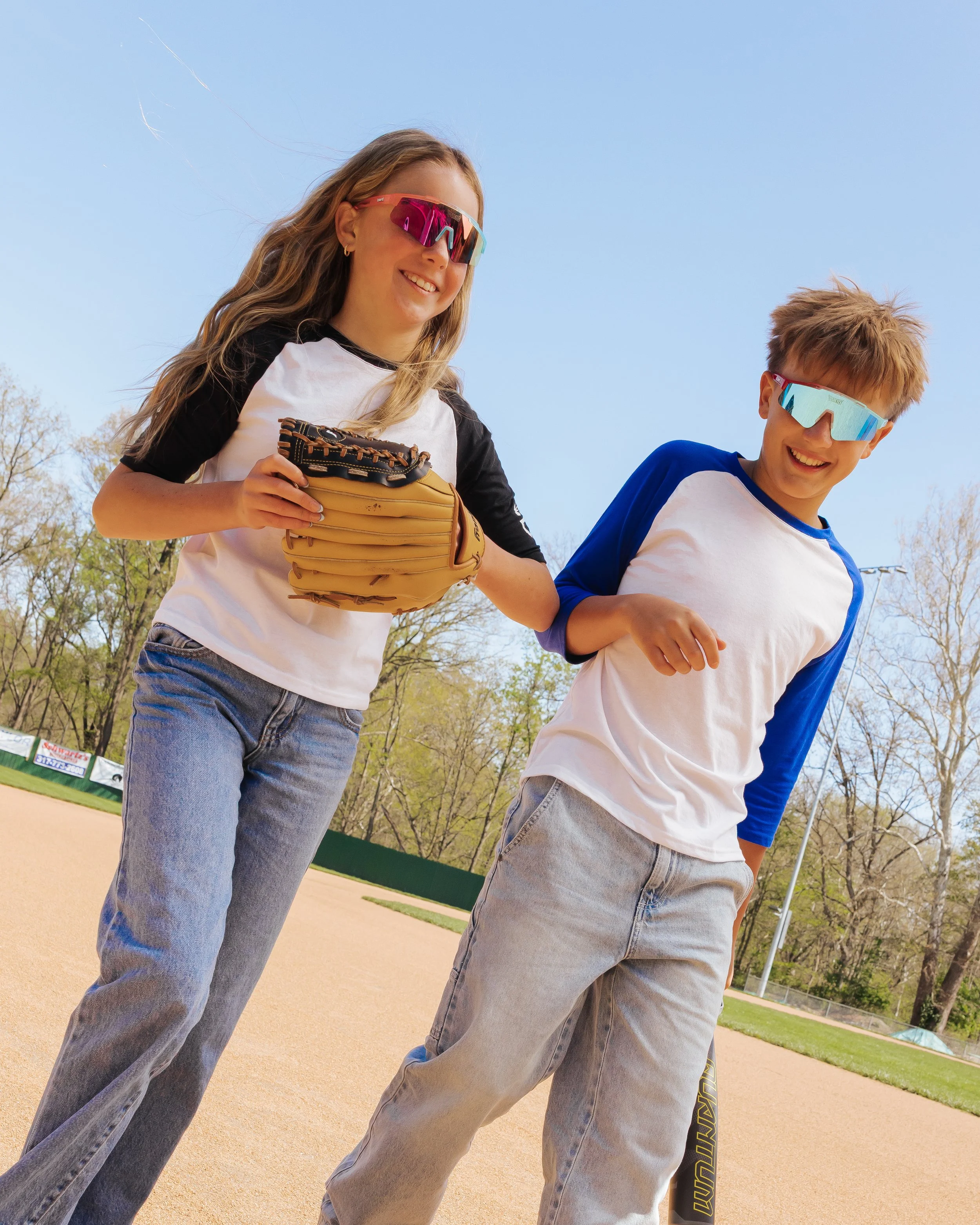 Two kids, a girl with long, wavy blonde hair wearing sunglasses and a baseball glove, and a boy with short, tousled hair and sunglasses, are smiling and standing outdoors on a sunny day at a baseball field, with trees and fencing in the background.