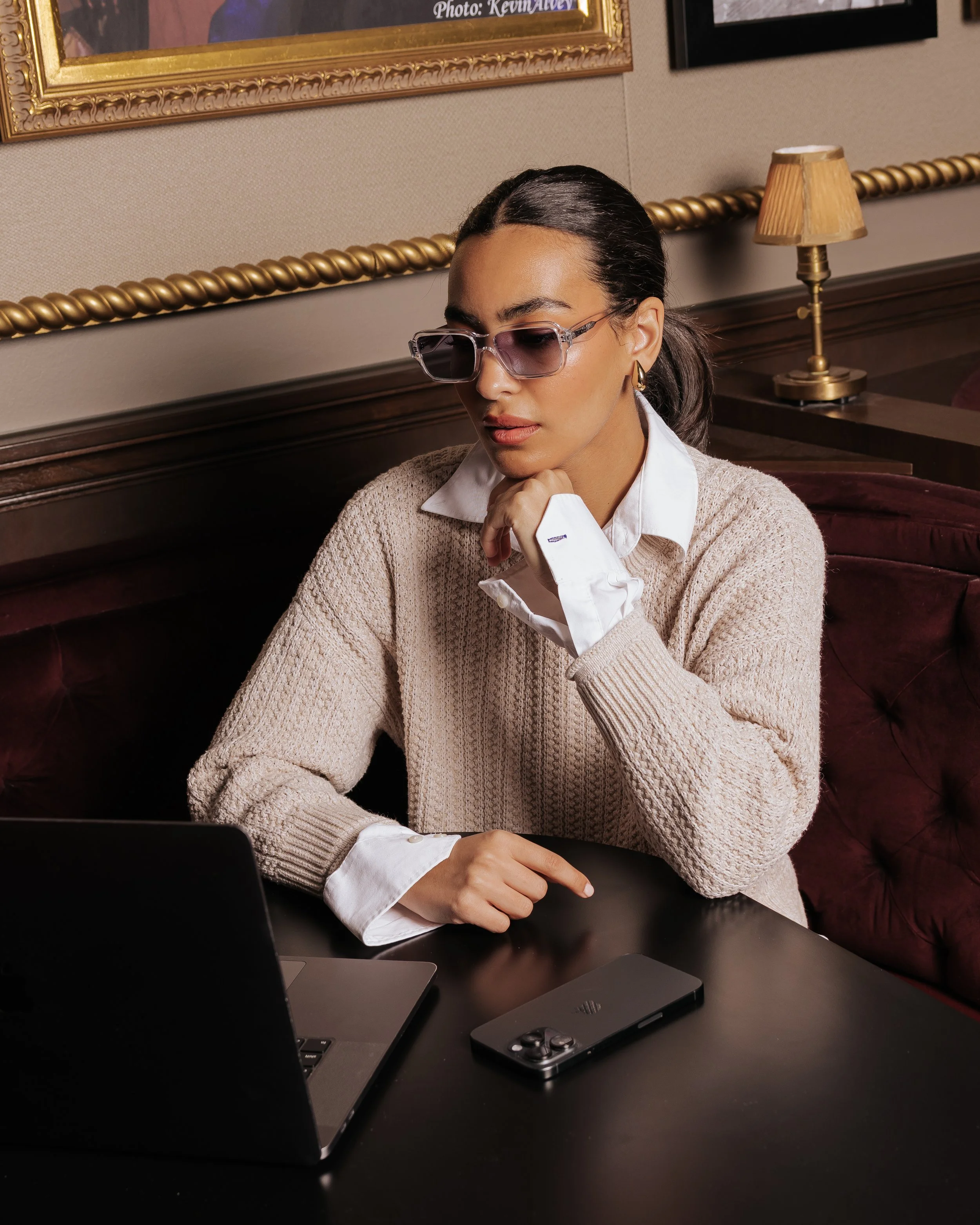 A woman with dark hair slicked back, wearing glasses, a white collared shirt, a beige sweater, gold hoop earrings, sitting at a black table with a laptop and a smartphone, in a room with beige walls, a gold-framed painting and a gold lamp.