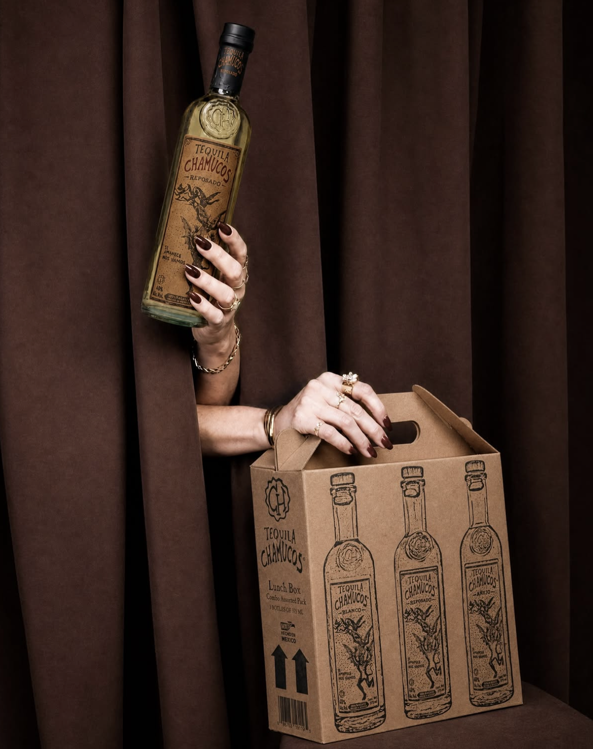 Person holding a bottle of Tequila Chamuco with brunette nails and jewelry, reaching into a cardboard box with similar bottles, against dark brown curtains.