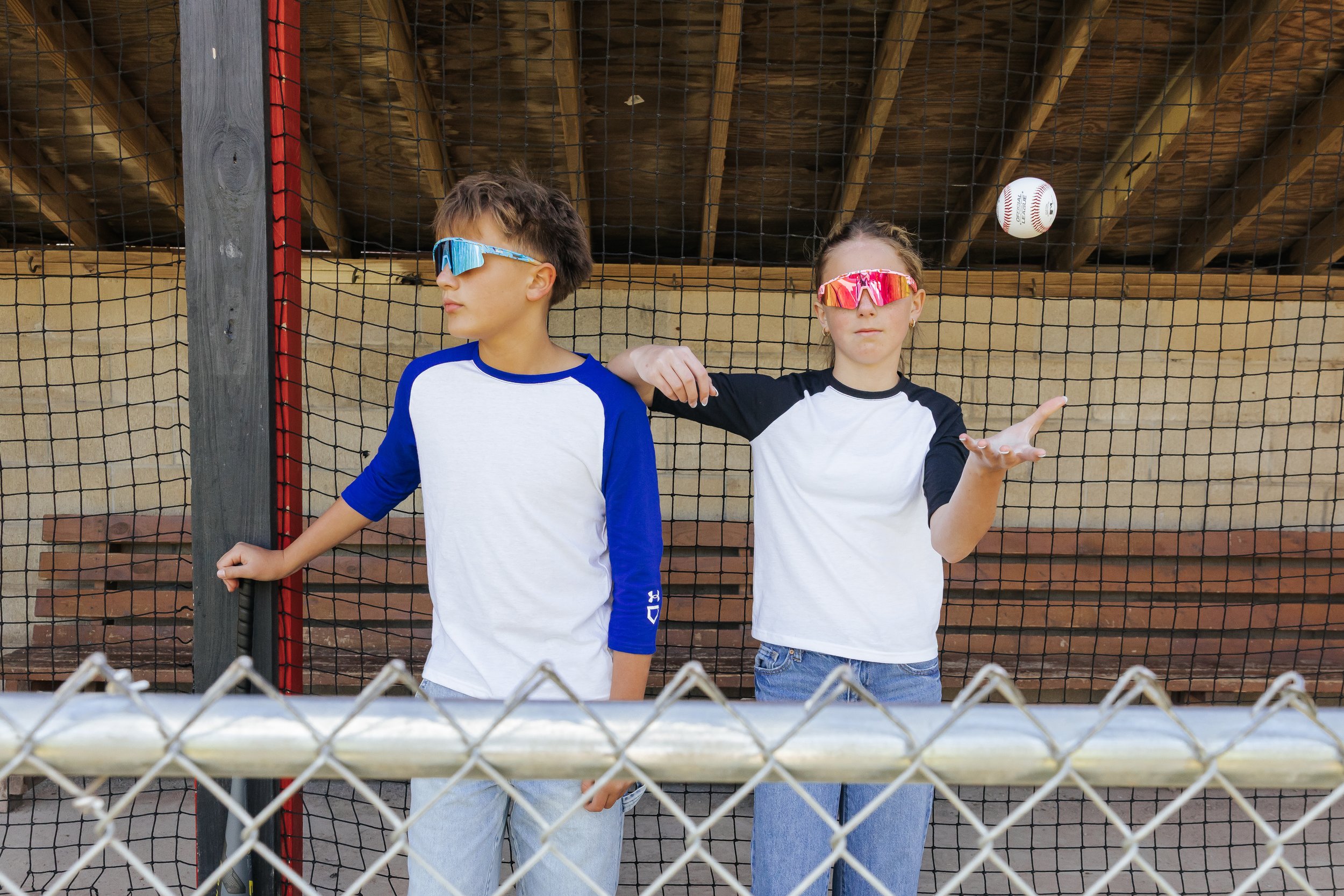 Two children wearing sports glasses and casual clothes stand behind a chain-link fence at a baseball field, with a baseball in mid-air above them.