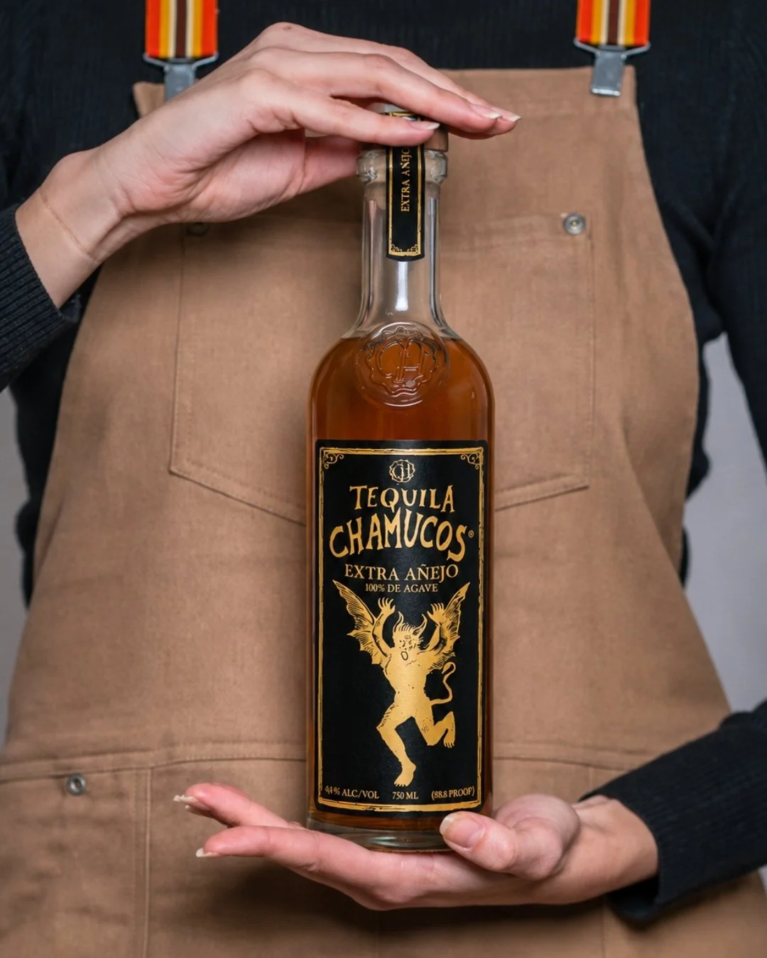 Person holding a bottle of tequila chamucos extra añejo with a black and gold label, showing a dragon-like creature, wearing a black shirt and brown apron.