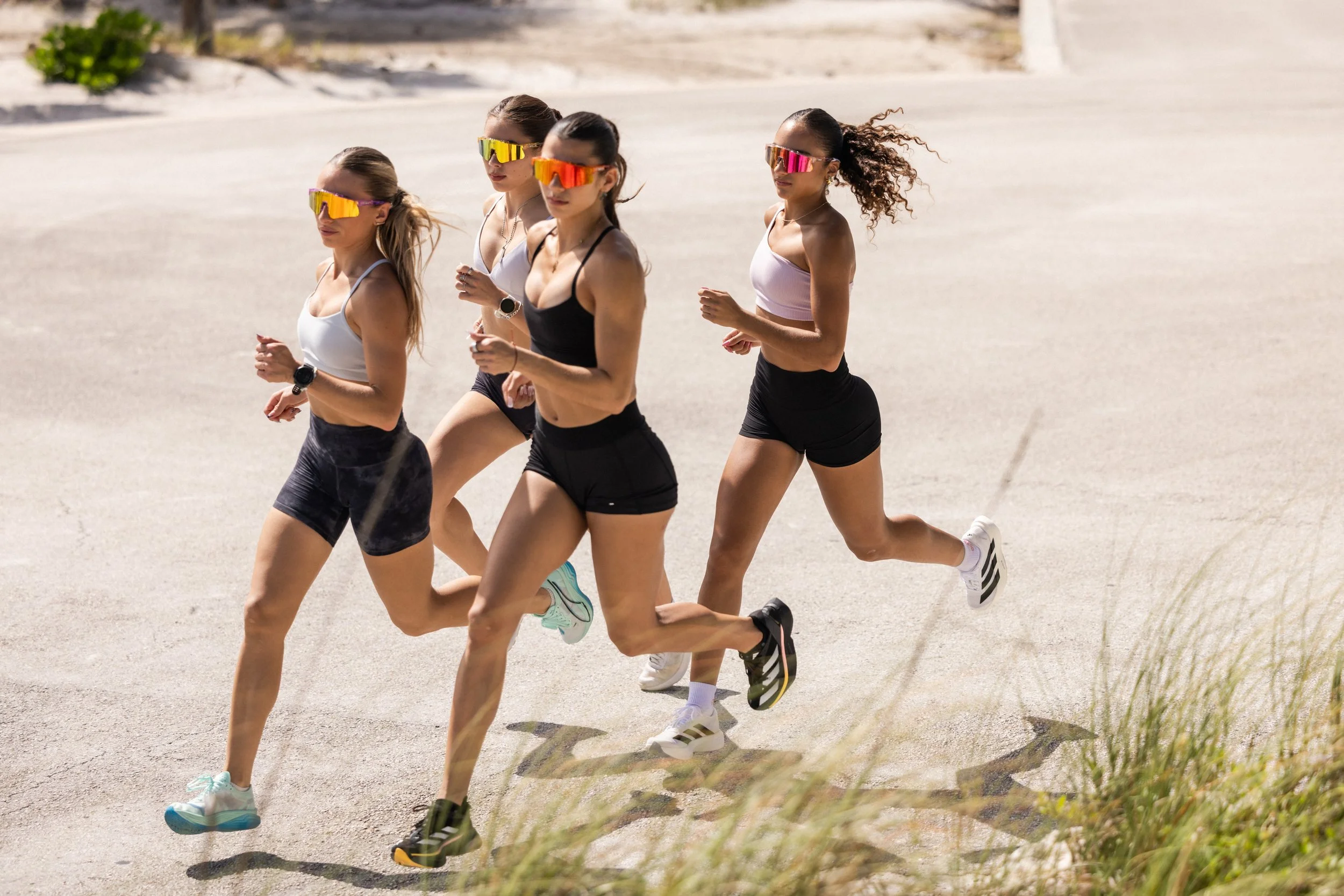 Four women running outdoors on a sunny day, wearing athletic clothing and colorful sunglasses.