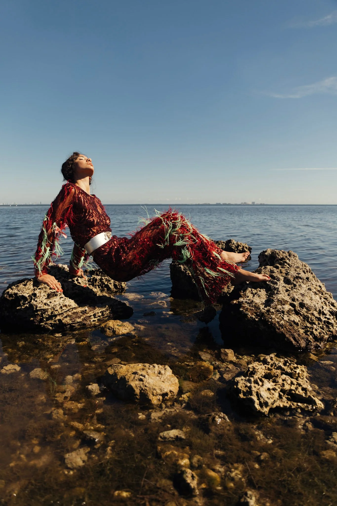 A woman in a red dress with floral patterns, sitting on rocks by the water with her eyes closed and face tilted upwards.