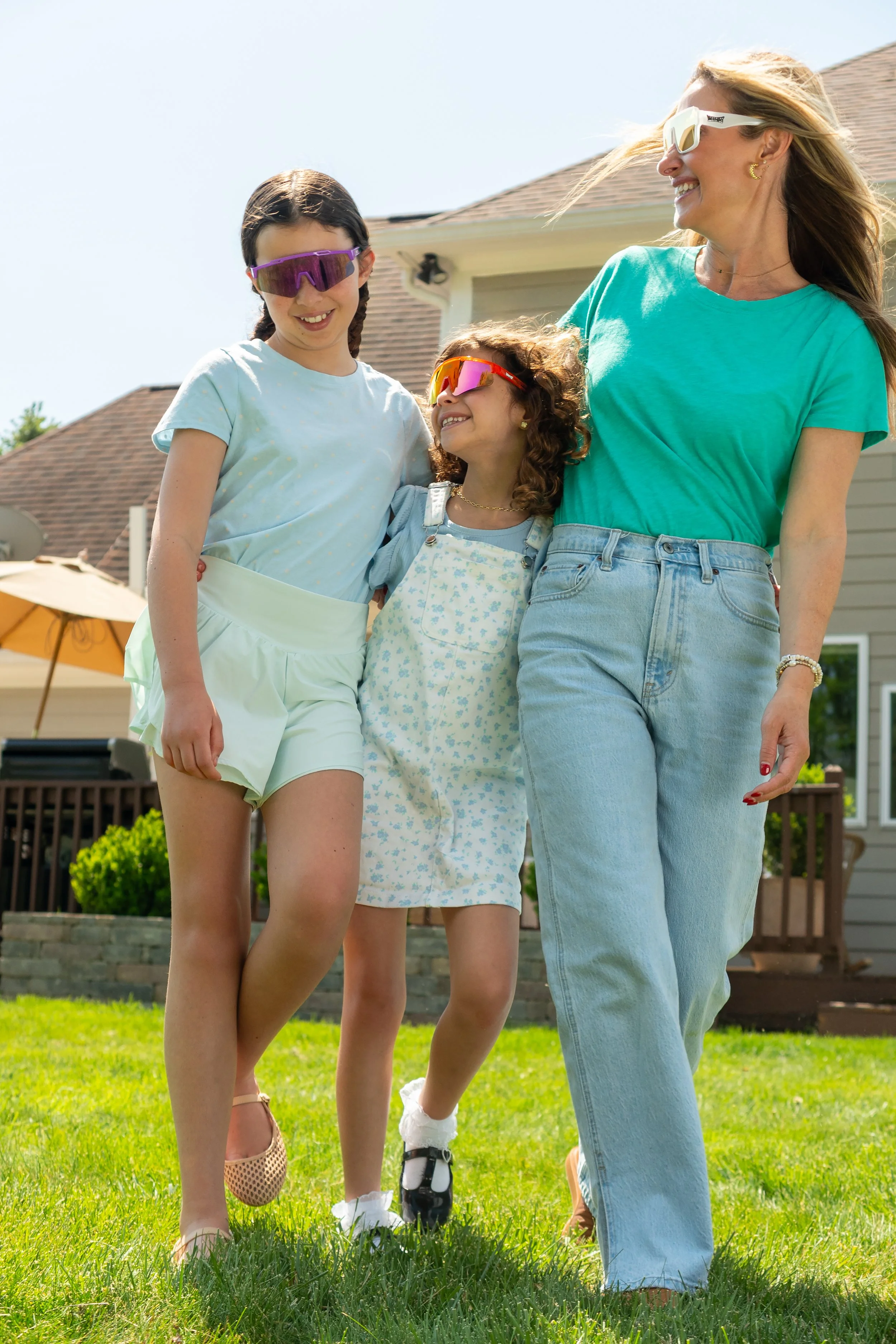 Woman and two girls walking on a lawn in a backyard, all wearing sunglasses and smiling, with a house, outdoor furniture, and umbrella in the background.