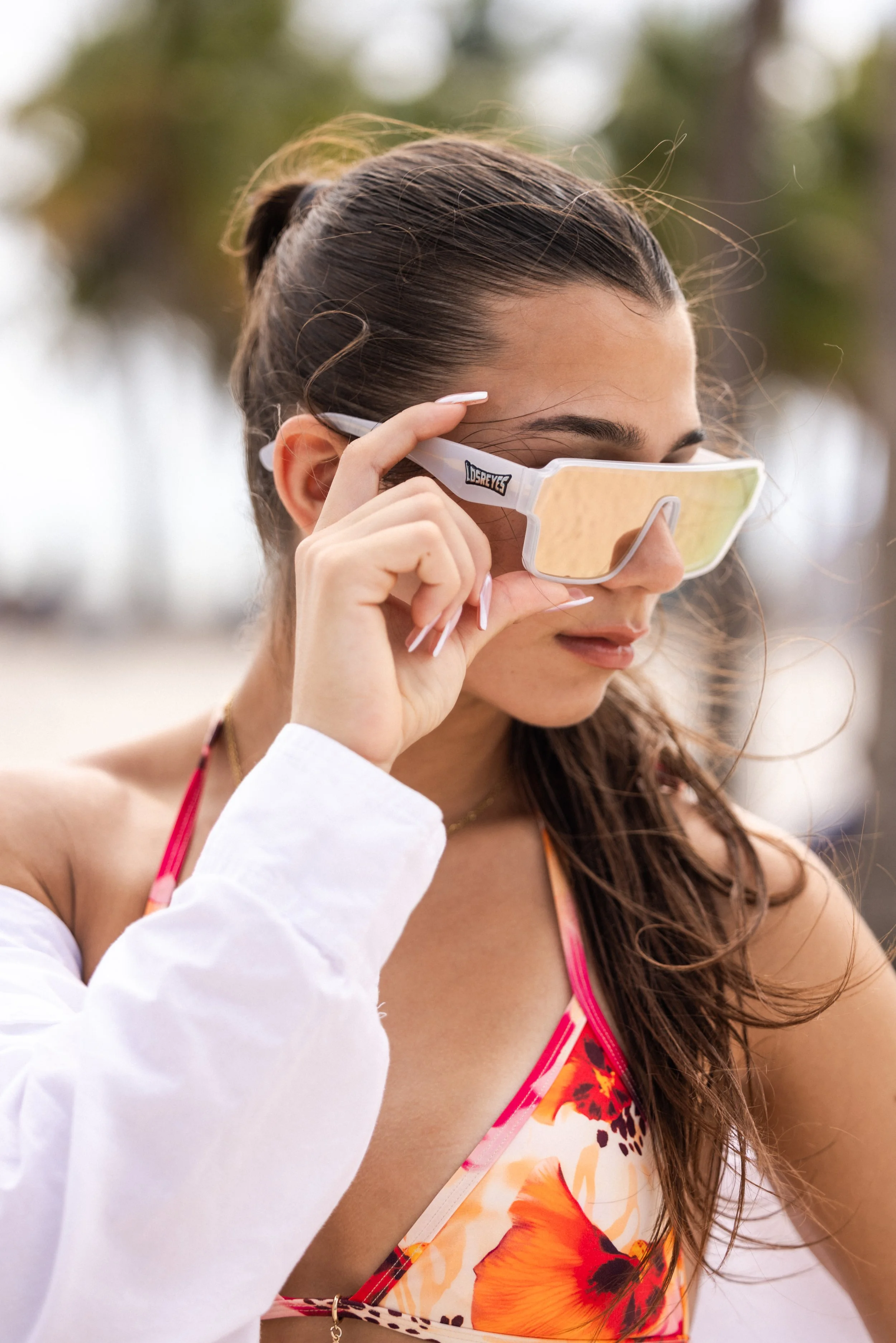 A woman with long dark hair tied back, wearing sunglasses and a colorful floral swimsuit with orange and red flowers, is outdoors. She is holding her sunglasses with her right hand, and her eyes are closed as if enjoying the sun.