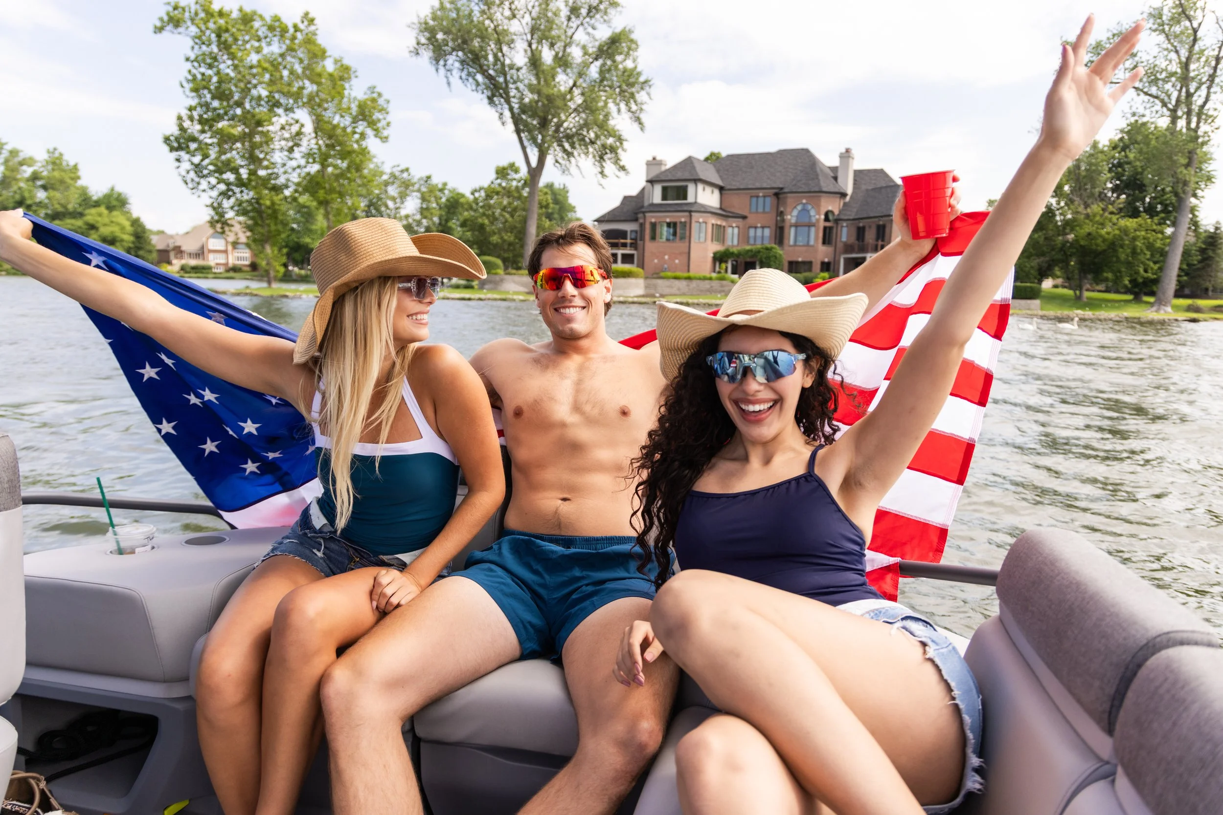 Group of friends on a boat, smiling, wearing summer clothing, hats, and sunglasses, holding American flags and a red cup, with a house and green trees in the background.
