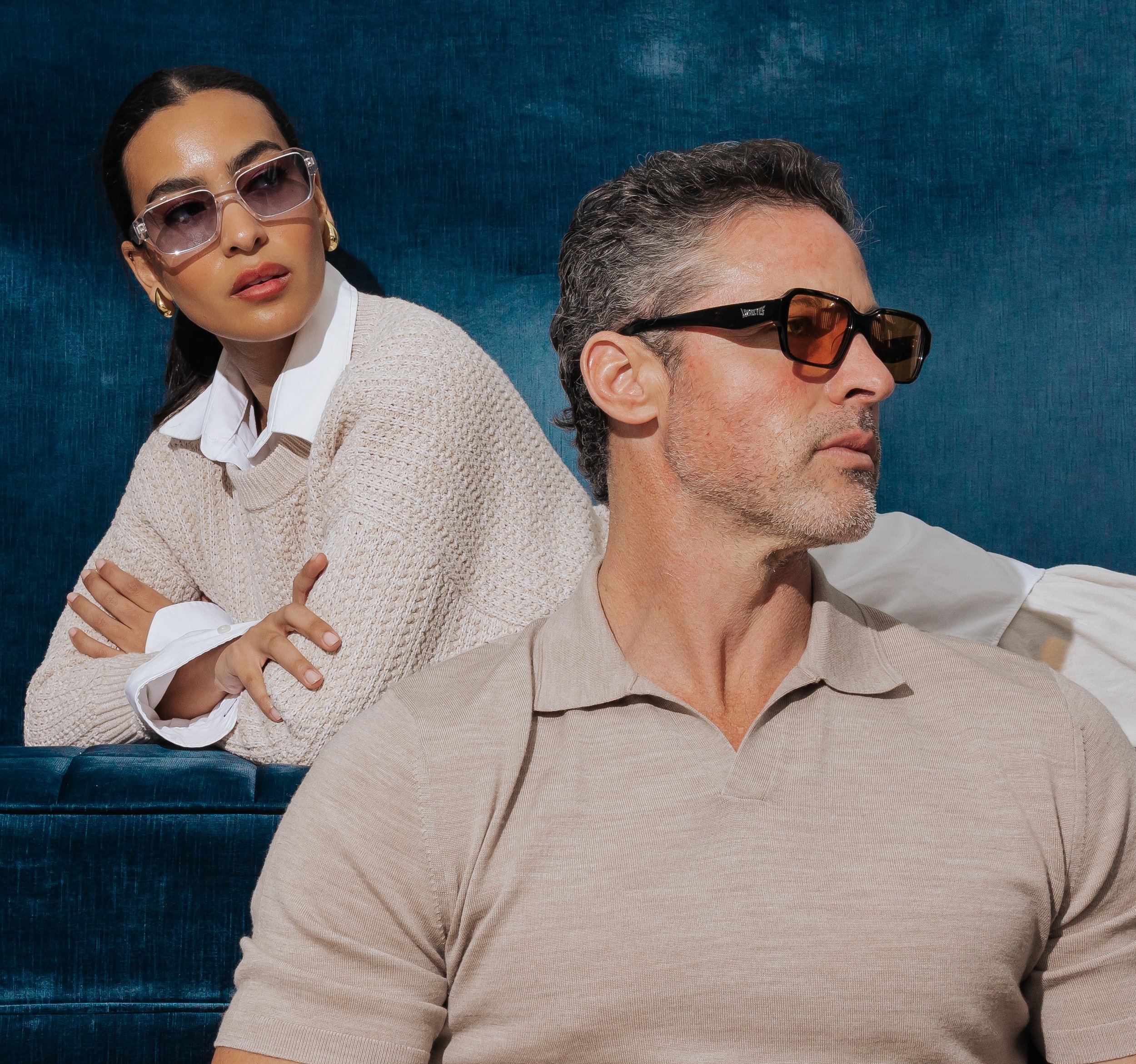 A man and a woman wearing sunglasses, sitting against a blue textured background. The woman is positioned behind the man with her arms crossed over his shoulder, and is wearing gold hoop earrings, a white shirt, and a beige sweater. The man has short, curly, salt-and-pepper hair and is wearing a beige shirt.