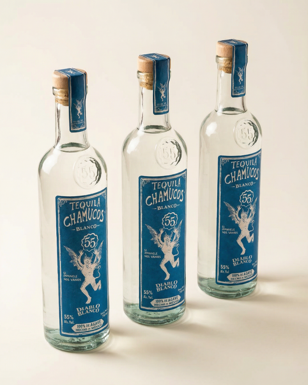 Three empty glass bottles of Tequila Chamuco Blanco with blue labels and cork stoppers, standing in a row on a white surface.