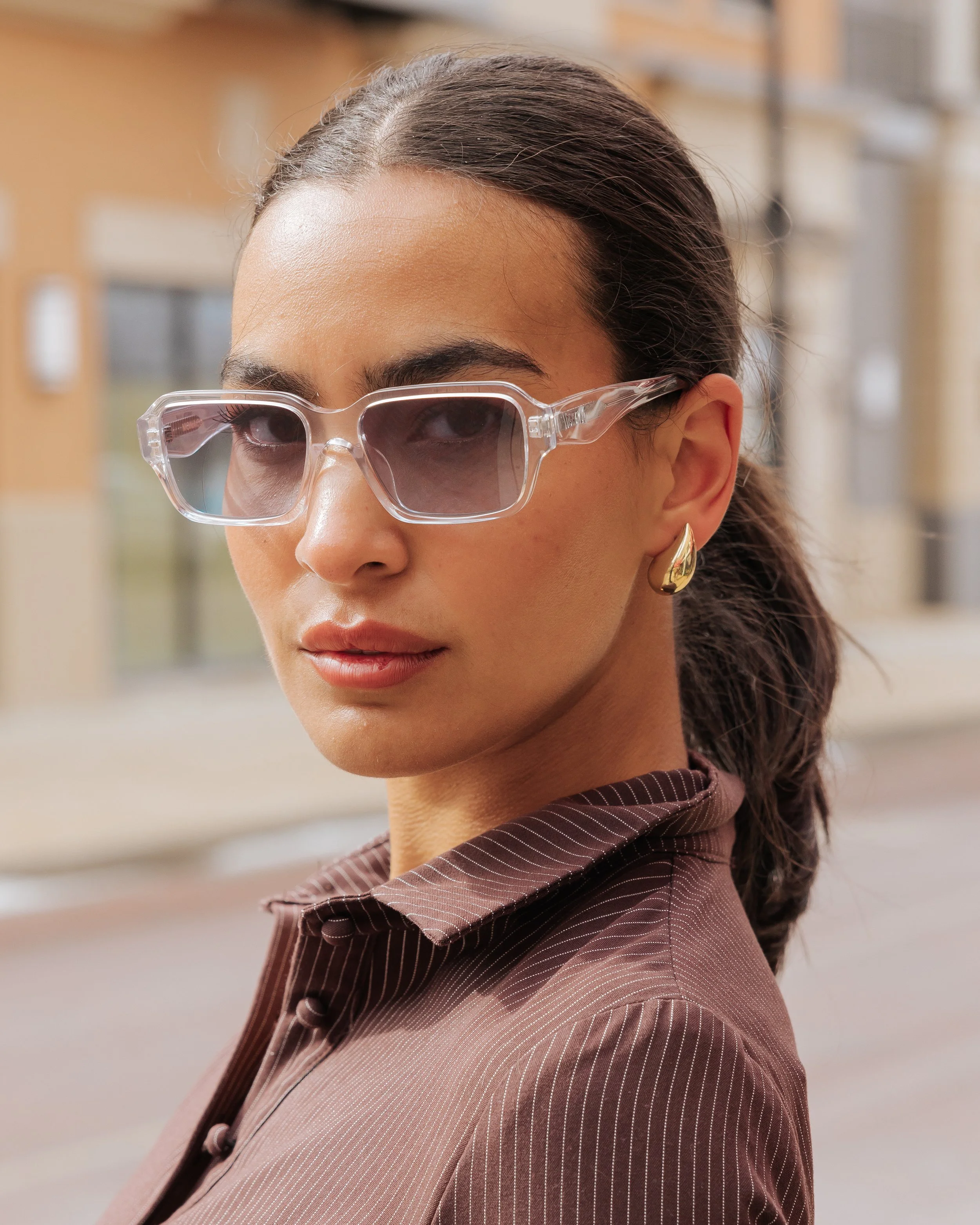 Close-up of a woman wearing clear, fashionable sunglasses, a brown striped shirt, and gold earrings, with her dark hair tied back, indoors.