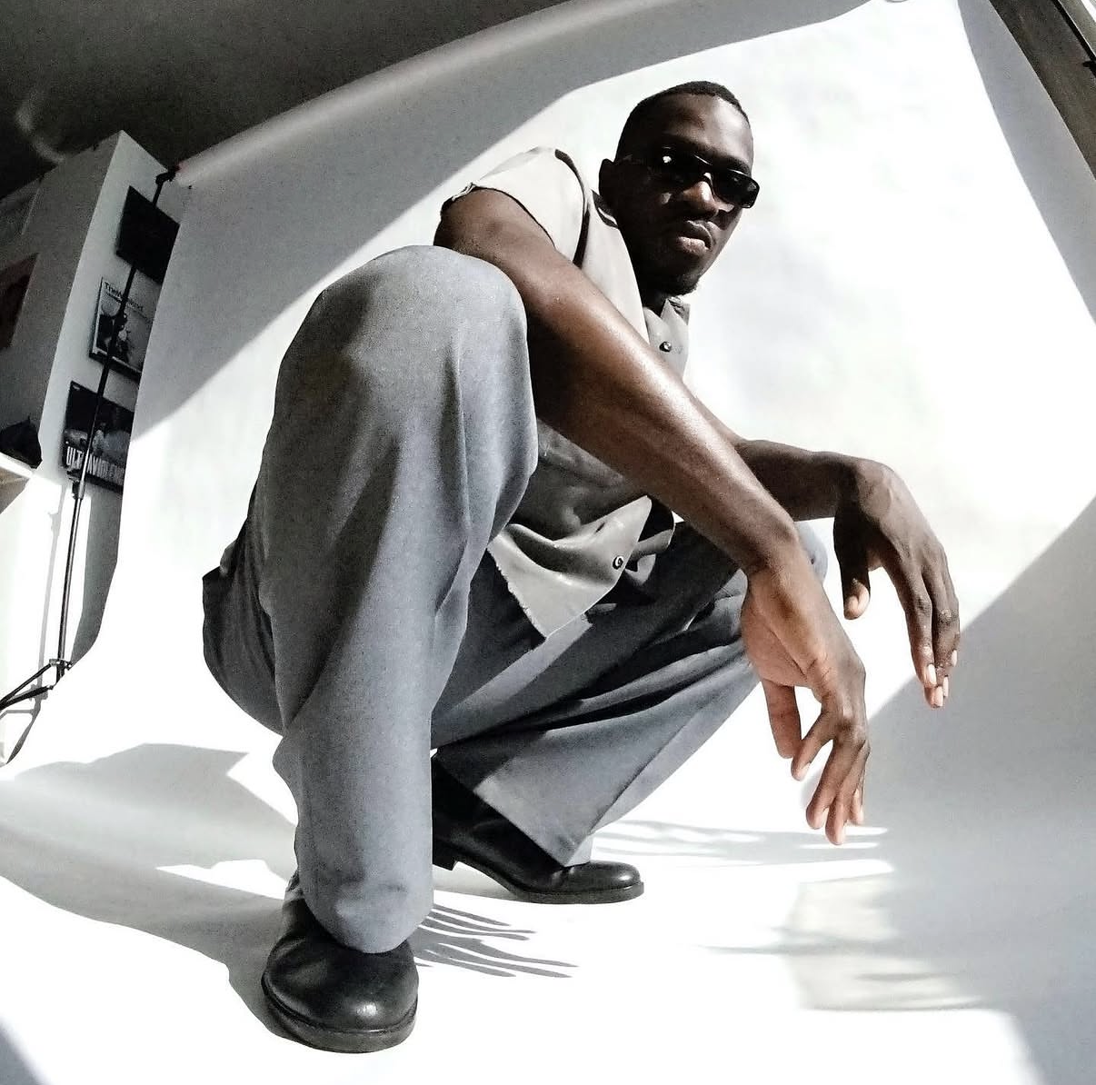 A man wearing sunglasses, a short-sleeved shirt, gray pants, and black shoes crouches in front of a white background in a photo studio.