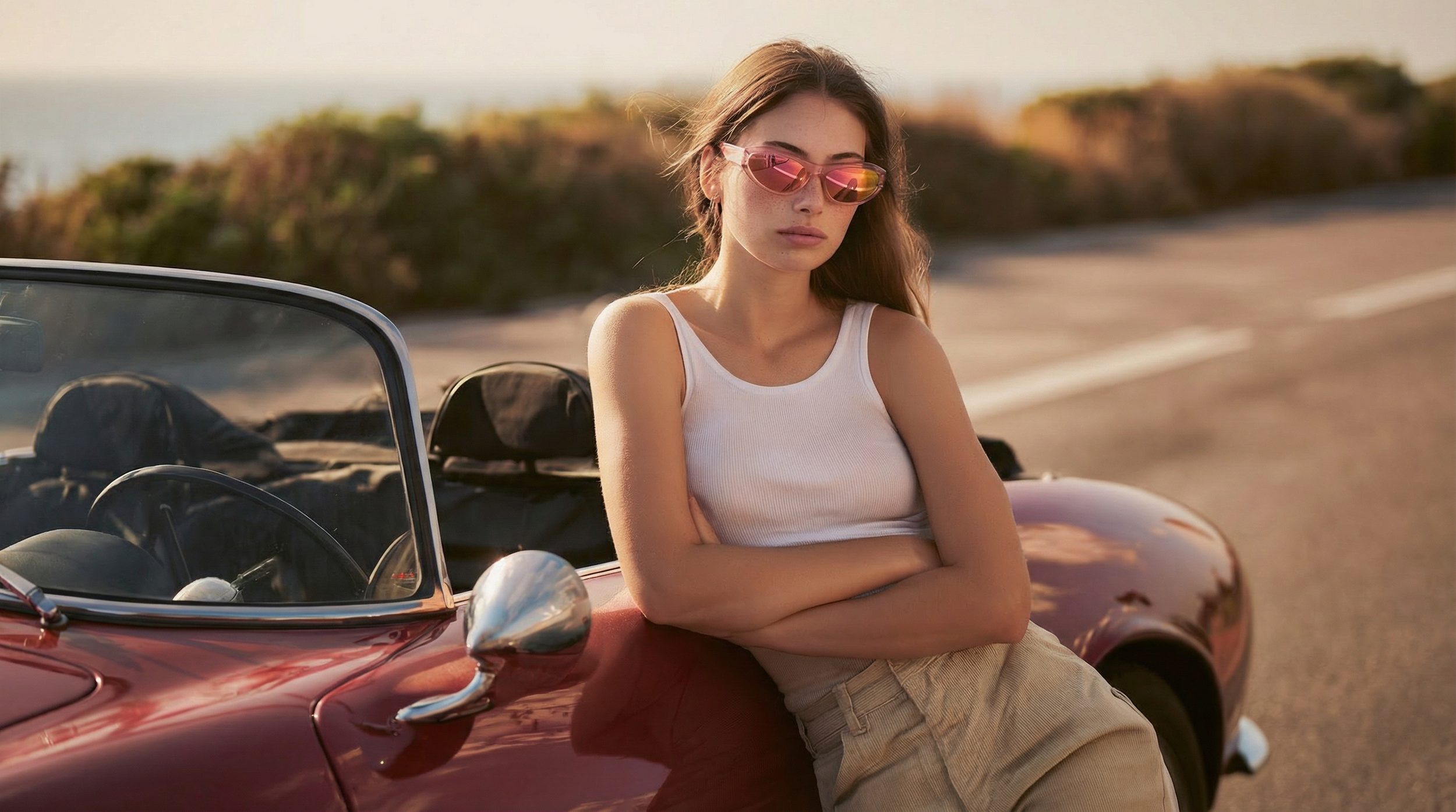 A young woman with long brown hair wearing pink reflective sunglasses, a white tank top, and beige pants, leaning against a vintage red convertible car on a road with a blurred landscape in the background.