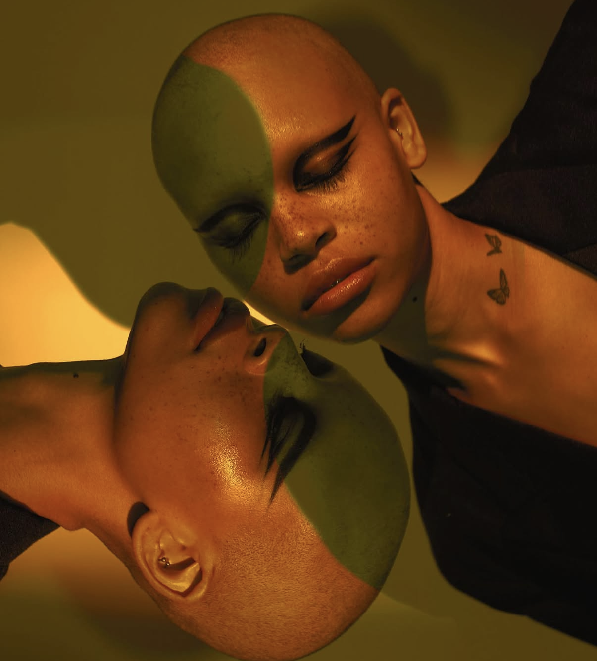 Two close-up portraits of a bald woman with face paint, including black eyeliner and freckles, against a yellow background. The woman has tattoos of butterflies and a small music note on her neck. In both images, the woman touches her face, with one eye closed and gentle expressions.