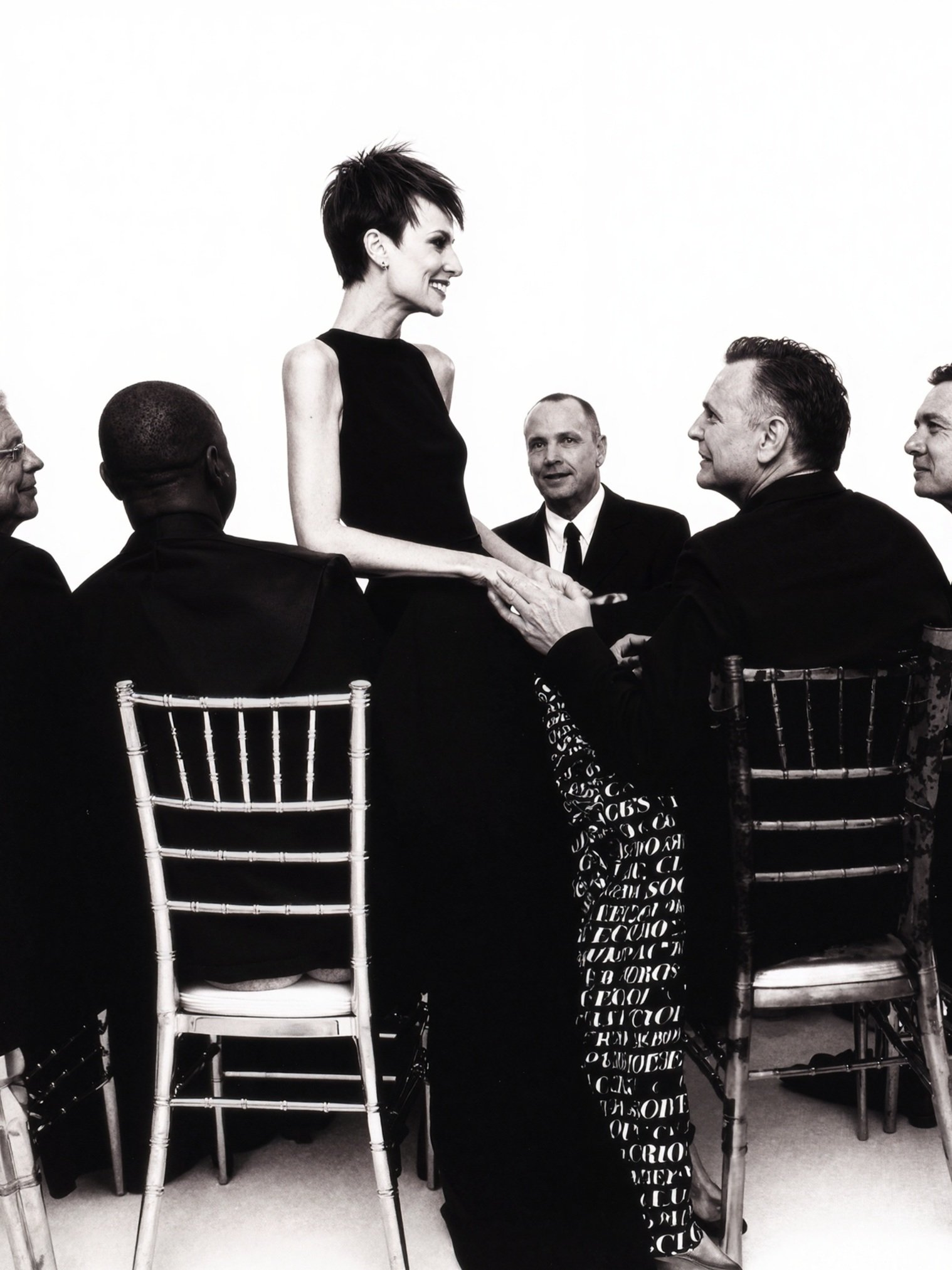 Black and white photo of a woman with a short spiky haircut smiling and holding hands of a man seated, surrounded by four other men sitting on chairs, all dressed in formal black suits, with some wearing glasses. They are in a well-lit room with a plain white background.