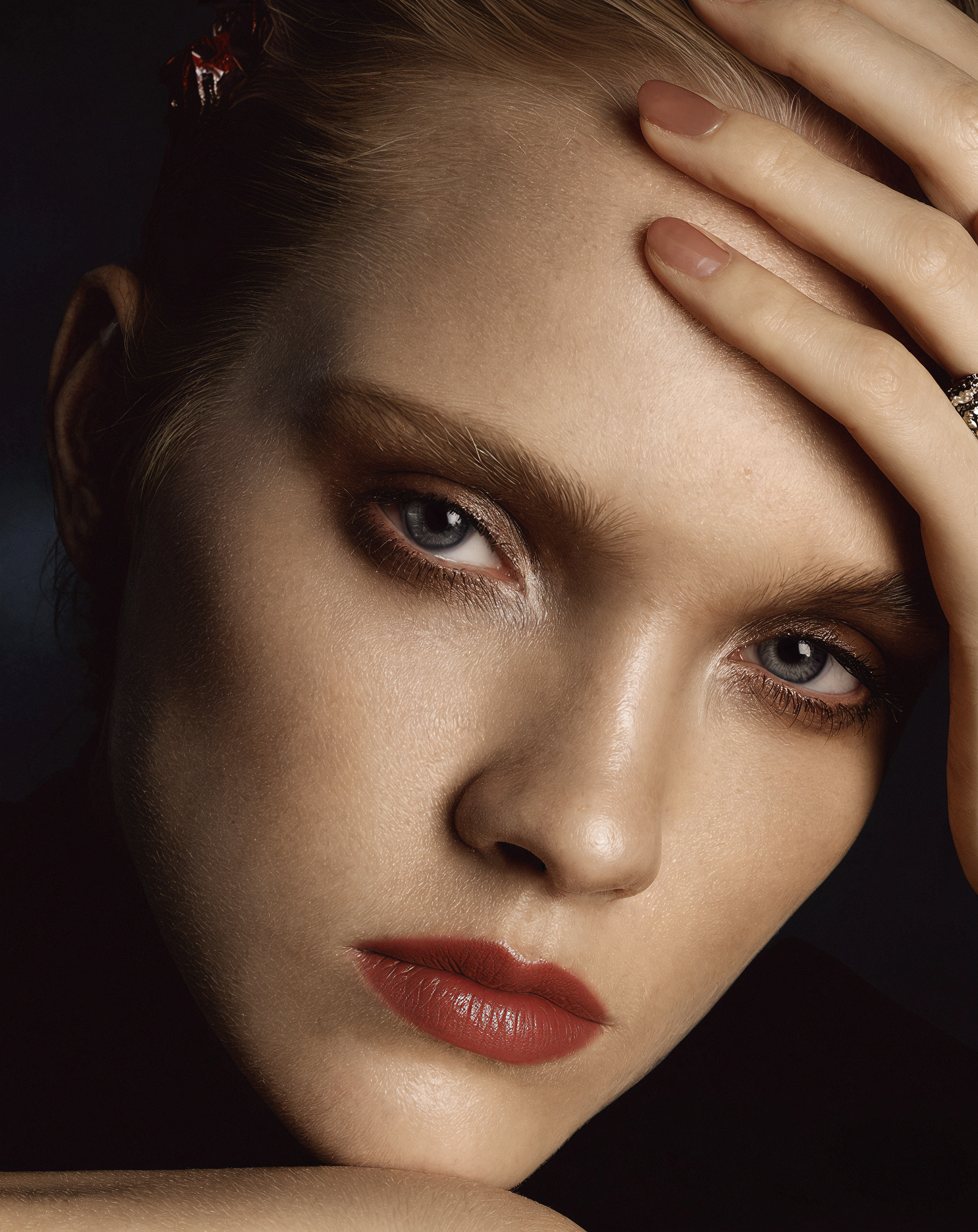Close-up of a woman's face with makeup, red lipstick, and her hand resting on her forehead.