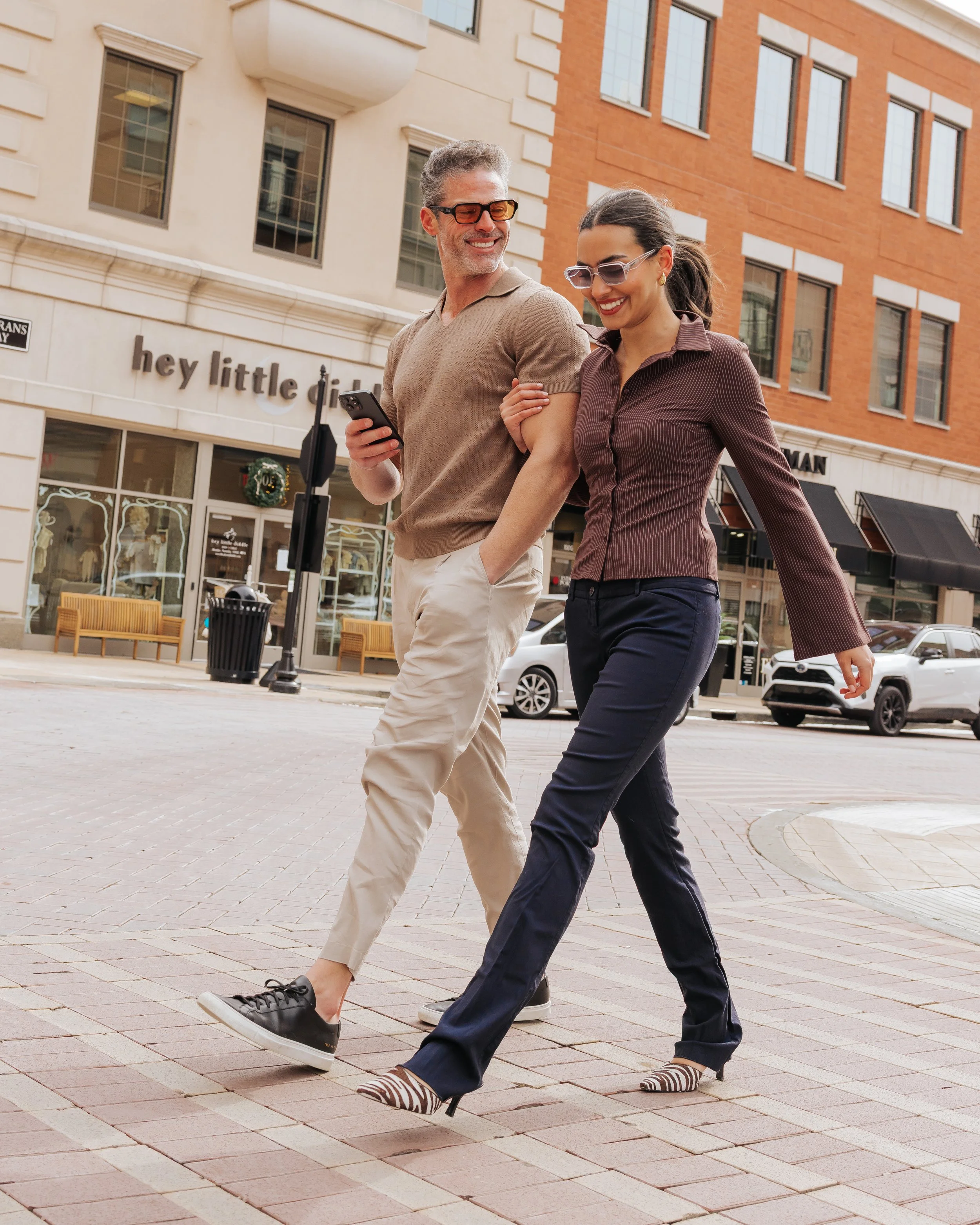 Two people, a man and a woman, walking together on a city street, smiling and looking at each other. The man is wearing beige pants, a beige shirt, black shoes, and sunglasses, and the woman is dressed in dark pants, a brown shirt, high heels, and sunglasses. They are enjoying a sunny day, and the background shows storefronts, parked cars, and brick buildings.