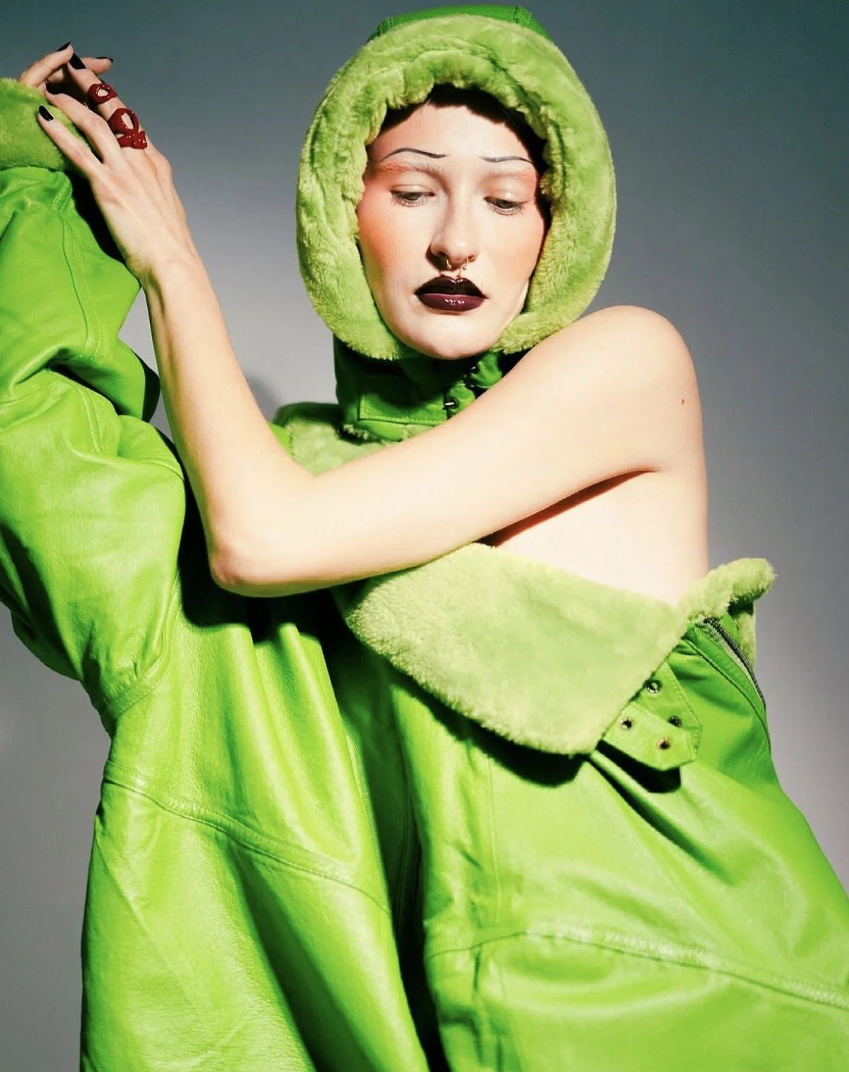 A woman with pale skin, dark lipstick, and a septum piercing wearing a bright green hooded jacket with faux fur lining, and matching gloves, posing with arms crossed and eyes lowered against a plain background.