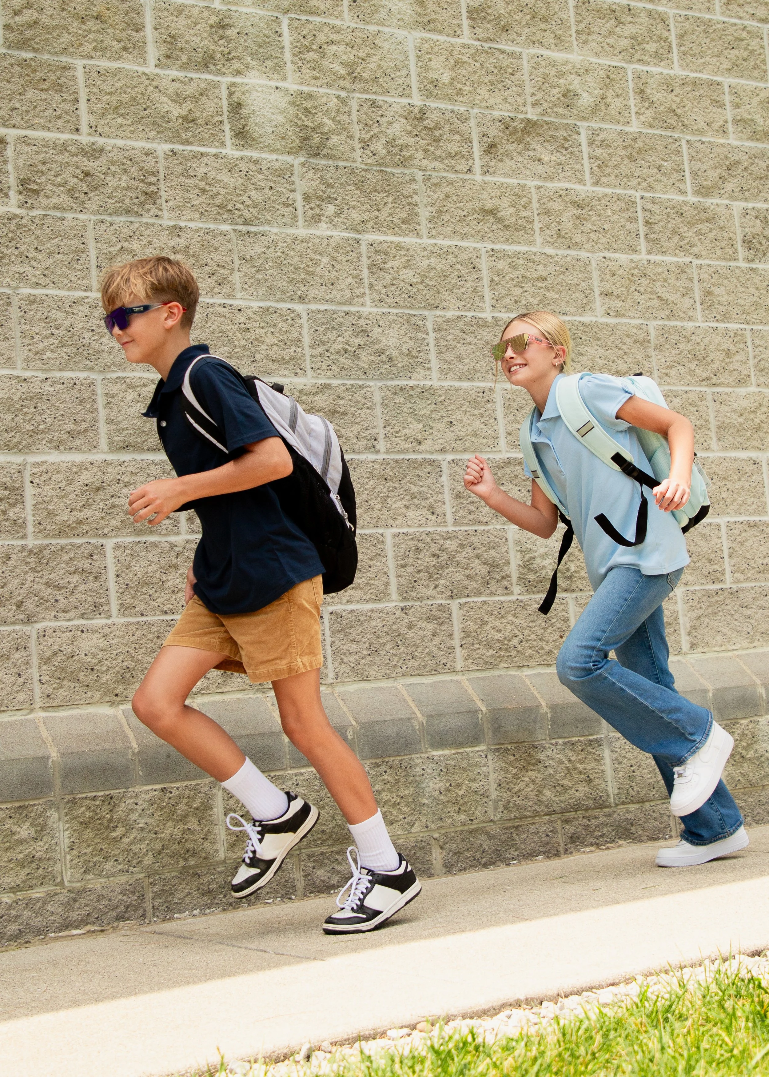 Two children, a boy and a girl, running along a sidewalk, wearing backpacks, sunglasses, and casual clothes, with a brick wall background.