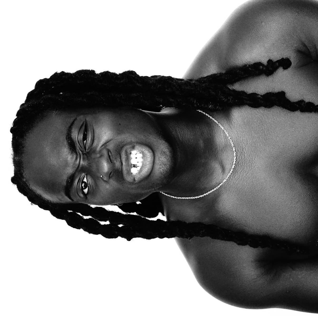 Black and white photo of a shirtless man with long dreadlocks, squinting and grimacing showing his teeth, wearing a thin chain necklace.
