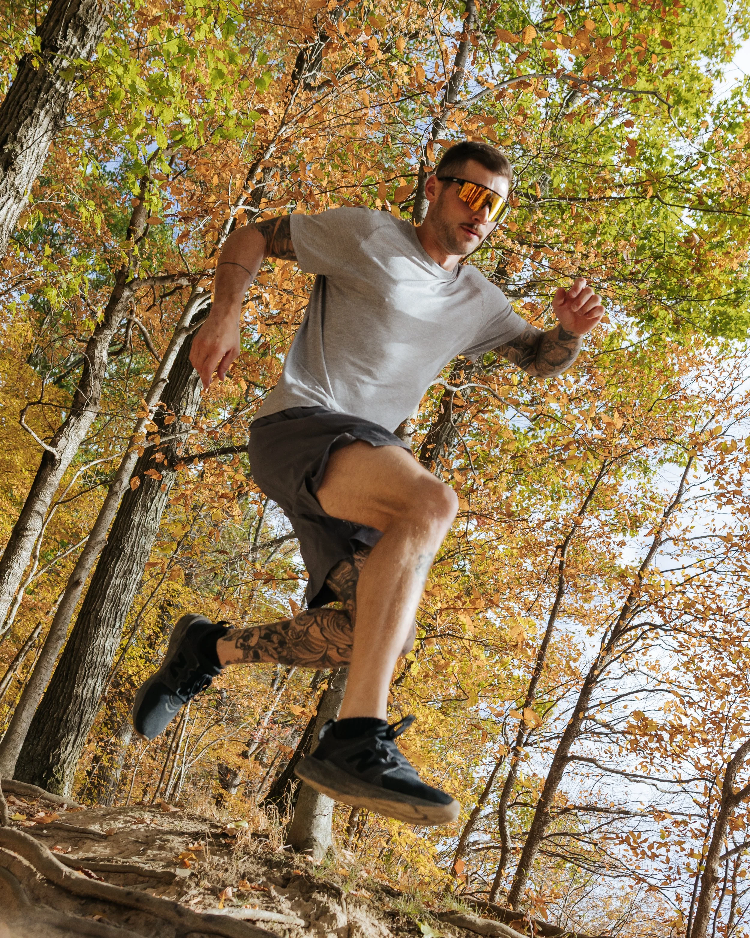 A man running outdoors among trees with autumn foliage, wearing sunglasses, a gray t-shirt, shorts, and sneakers, with a tattoo on his arm.