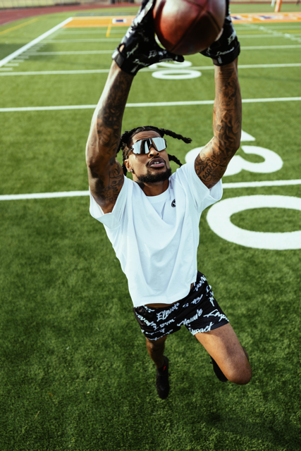 A male football player practicing on a football field, jumping with arms extended to catch or block a football overhead, wearing sunglasses, a white T-shirt, black shorts with white writing, and black gloves.