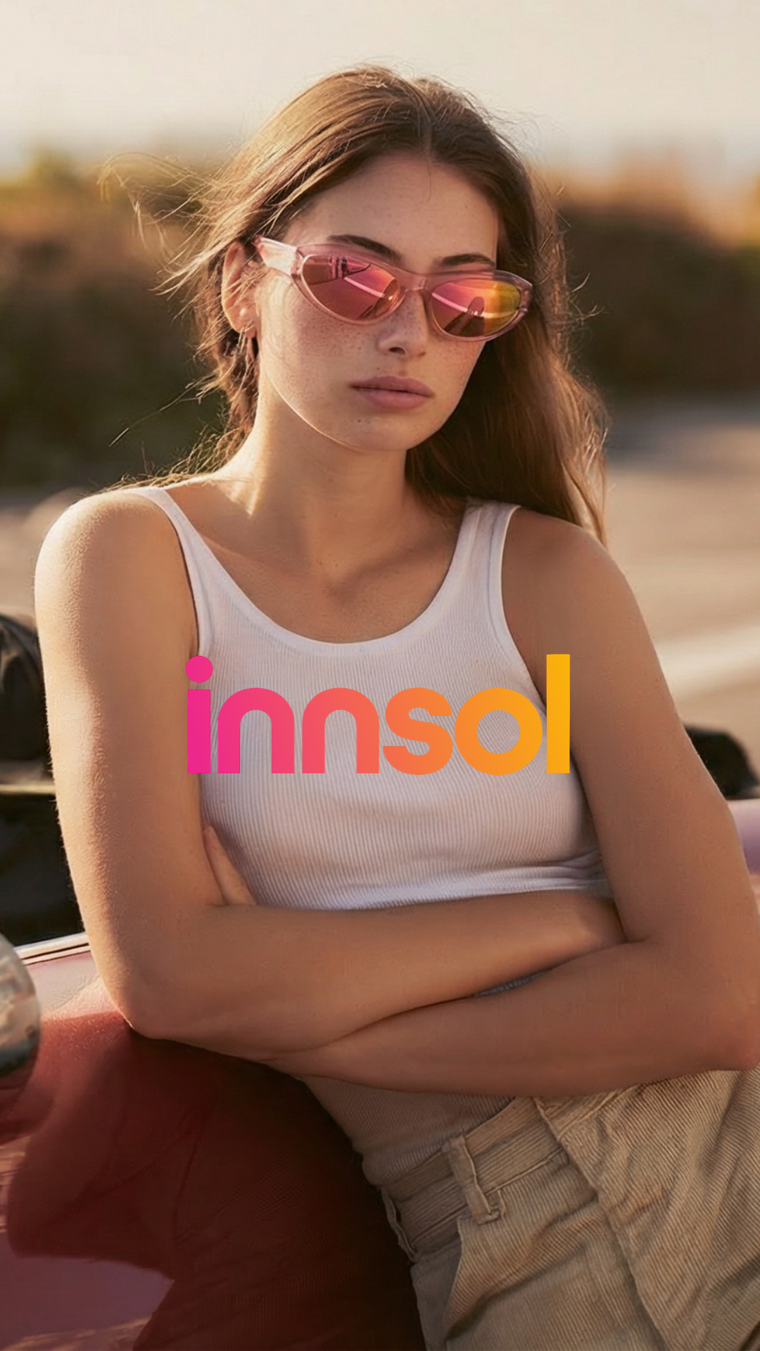 A young woman with long brown hair, wearing pink sunglasses and a white tank top, sitting outdoors with arms crossed, with the word "inns
