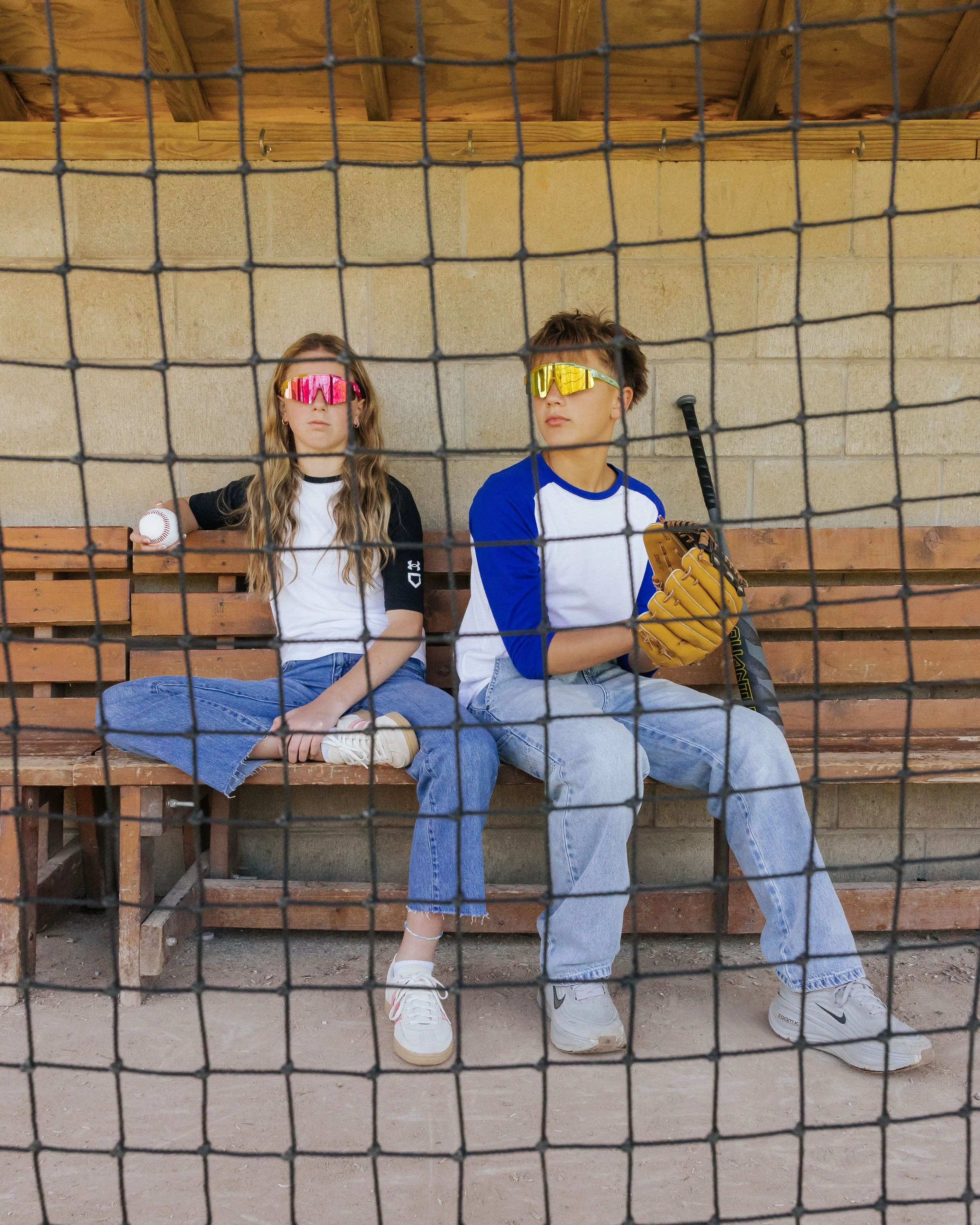 Two young baseball players sitting on a wooden bench inside a dugout behind a chain-link fence. The girl on the left has long wavy hair and is wearing pink reflective sports sunglasses, a white and black baseball jersey, blue jeans, and white sneakers, and is holding a baseball. The boy on the right has short hair and is wearing yellow reflective sports sunglasses, a white and blue baseball jersey, light blue jeans, and white Nike sneakers, holding a baseball bat and wearing a yellow baseball glove.