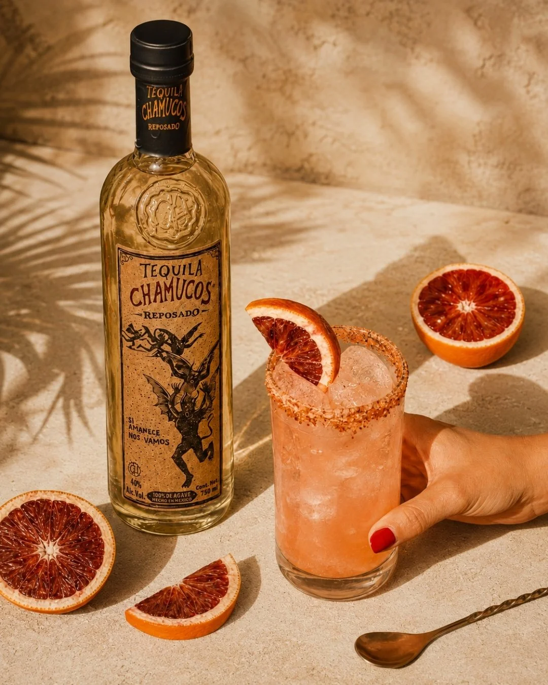 A bottle of Chamucos Reposado tequila, a cocktail with blood orange slices and salted rim, and sliced blood oranges on a beige surface.