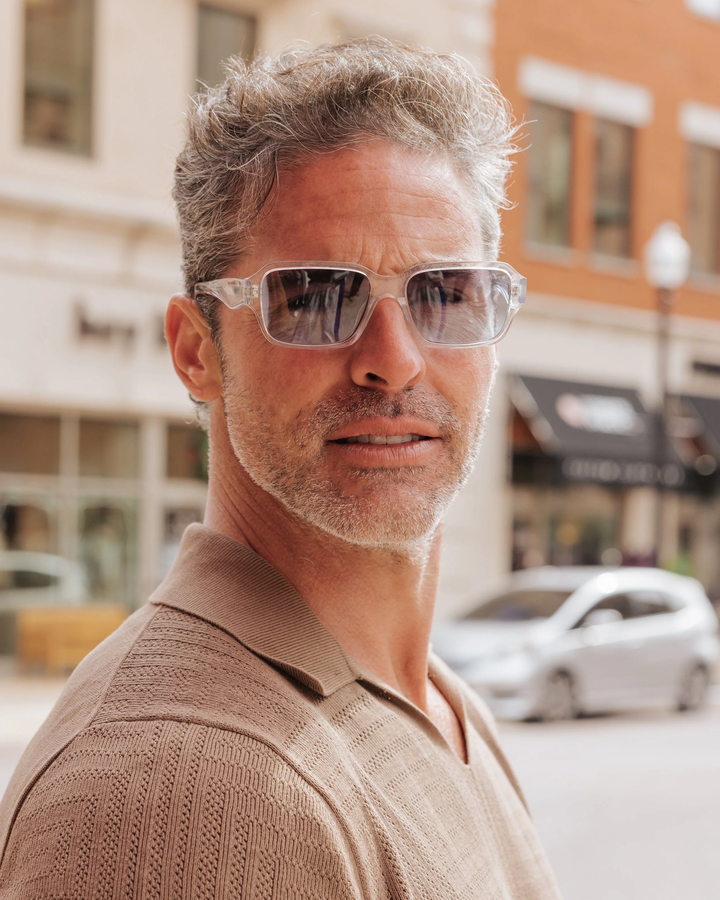Close-up of a middle-aged man with gray, curly hair wearing clear-framed sunglasses and a beige textured polo shirt on a city street.