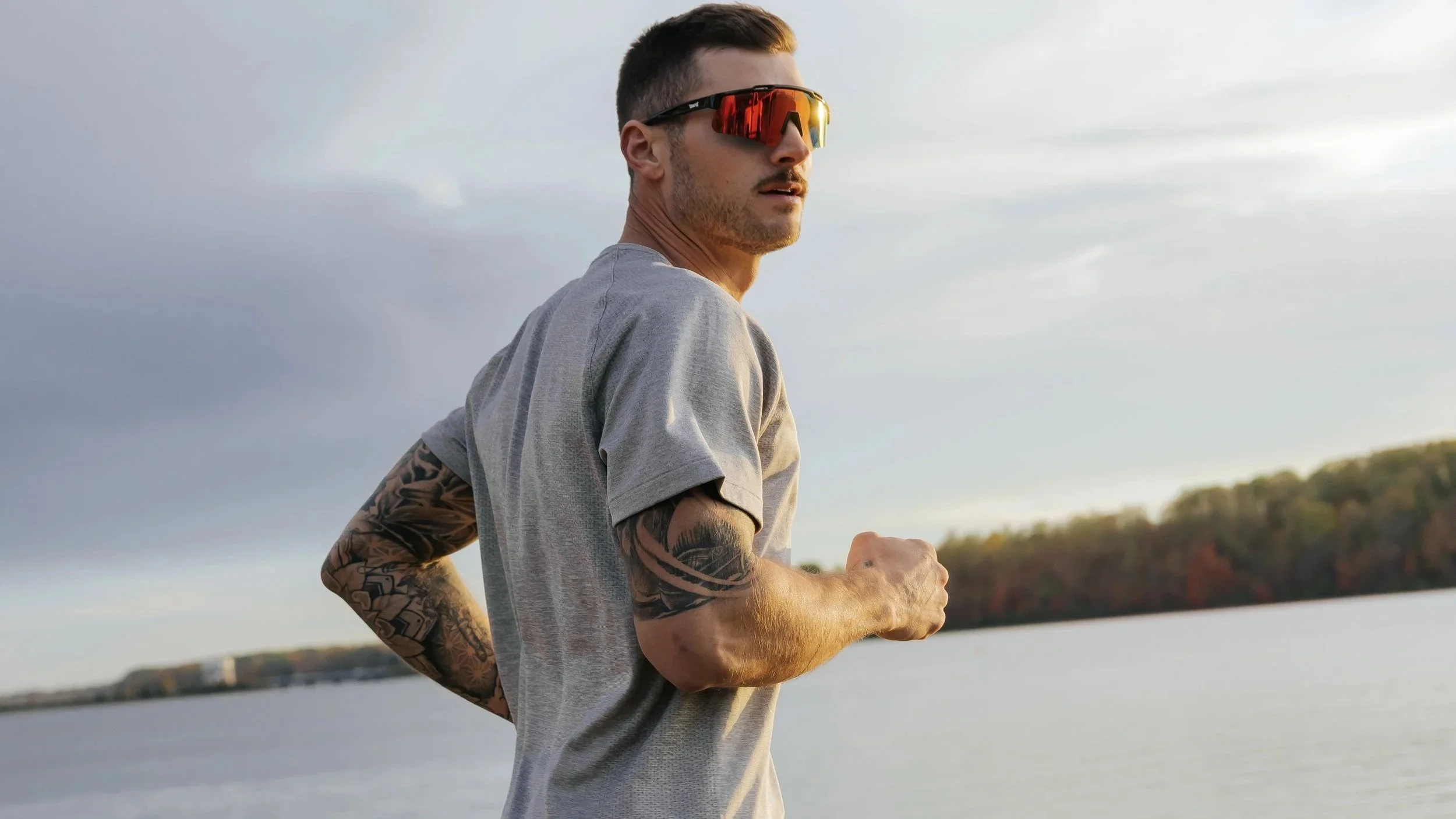 A young man jogging outdoors near a body of water, wearing sunglasses and a gray t-shirt, with tattoos on his arms, during sunset or sunrise.