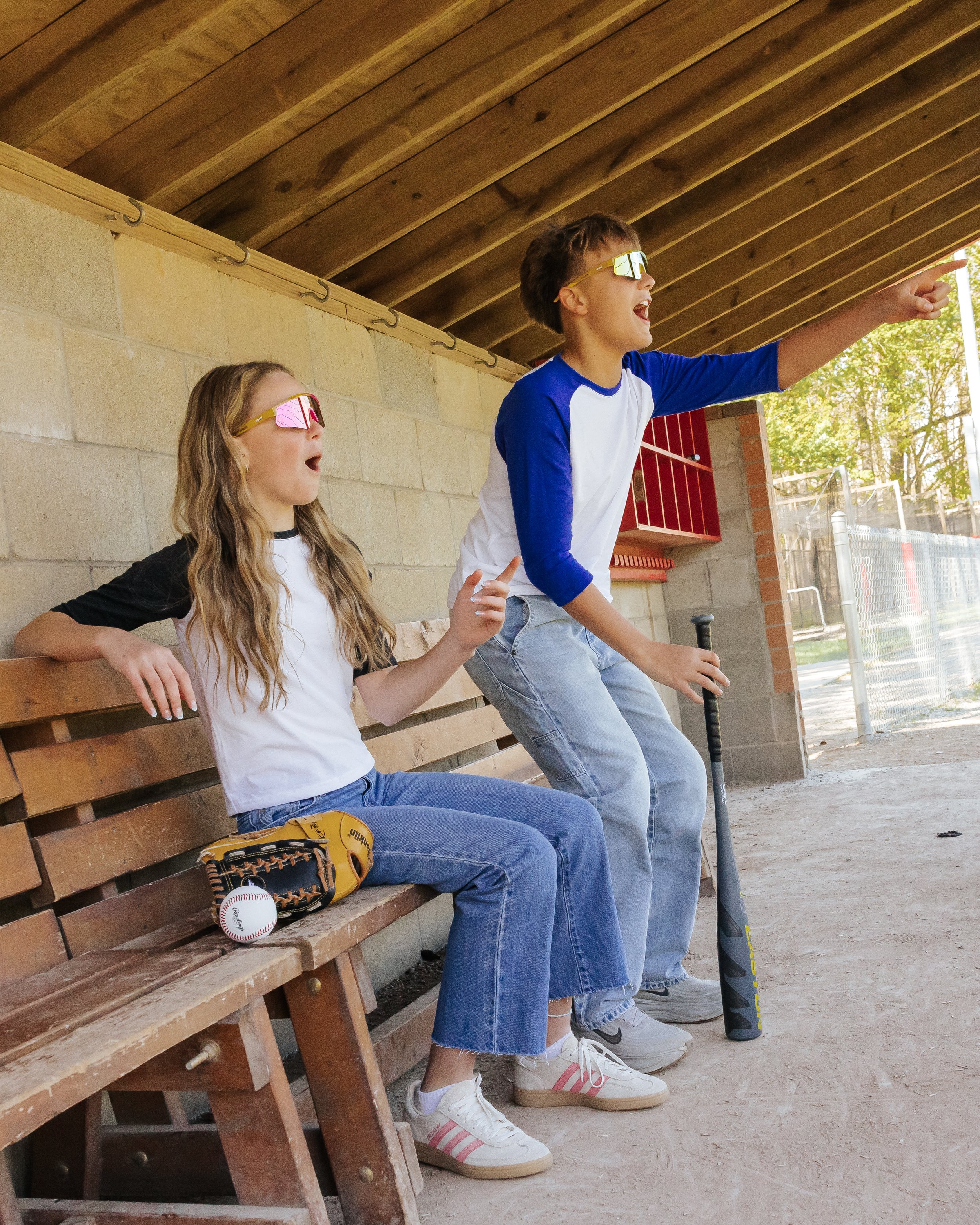 Two kids sitting on a wooden bench at a baseball field, wearing sunglasses and sports gear, one pointing and the other holding a baseball bat, with baseball equipment and dugout in the background.