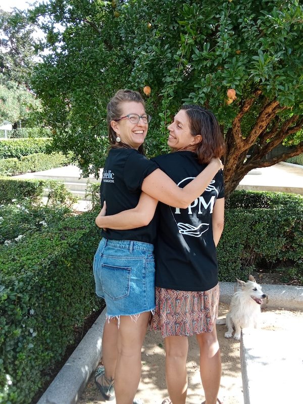 Two women wearing Psychedelic Pathways Magazine T-shirts embrace and smile at each other outdoors beneath a pomegranate tree, with a small dog beside them.
