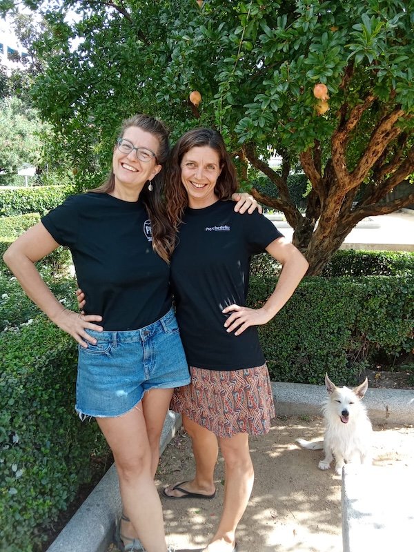Two women wearing Psychedelic Pathways Magazine T-shirts pose together outdoors beneath a pomegranate tree, smiling at the camera, with a small dog beside them.