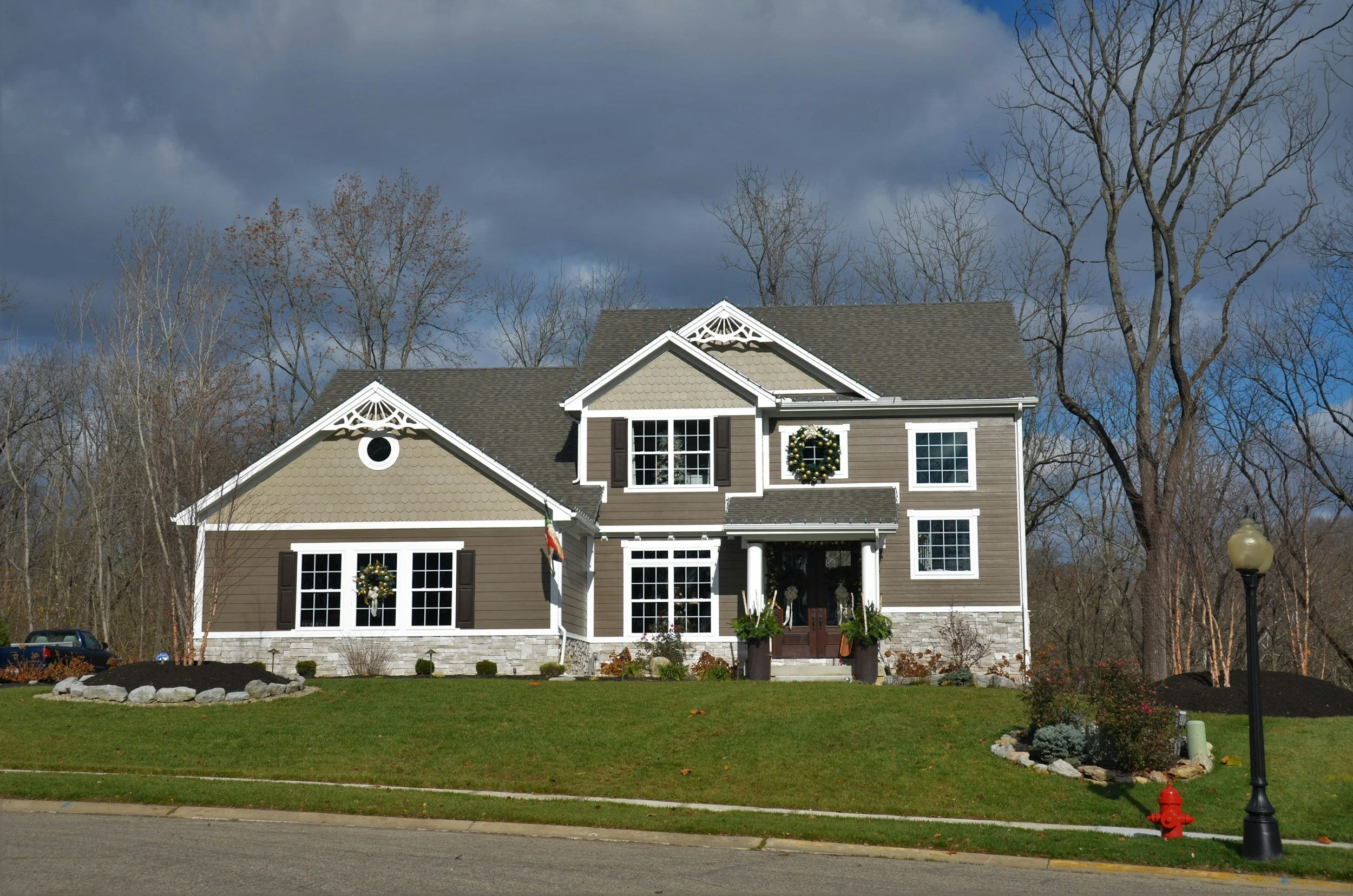 A two-story house decorated for Christmas with wreaths on the windows, a front porch, a flower bed, and a green lawn. There are leafless trees in the background and dark, cloudy sky.