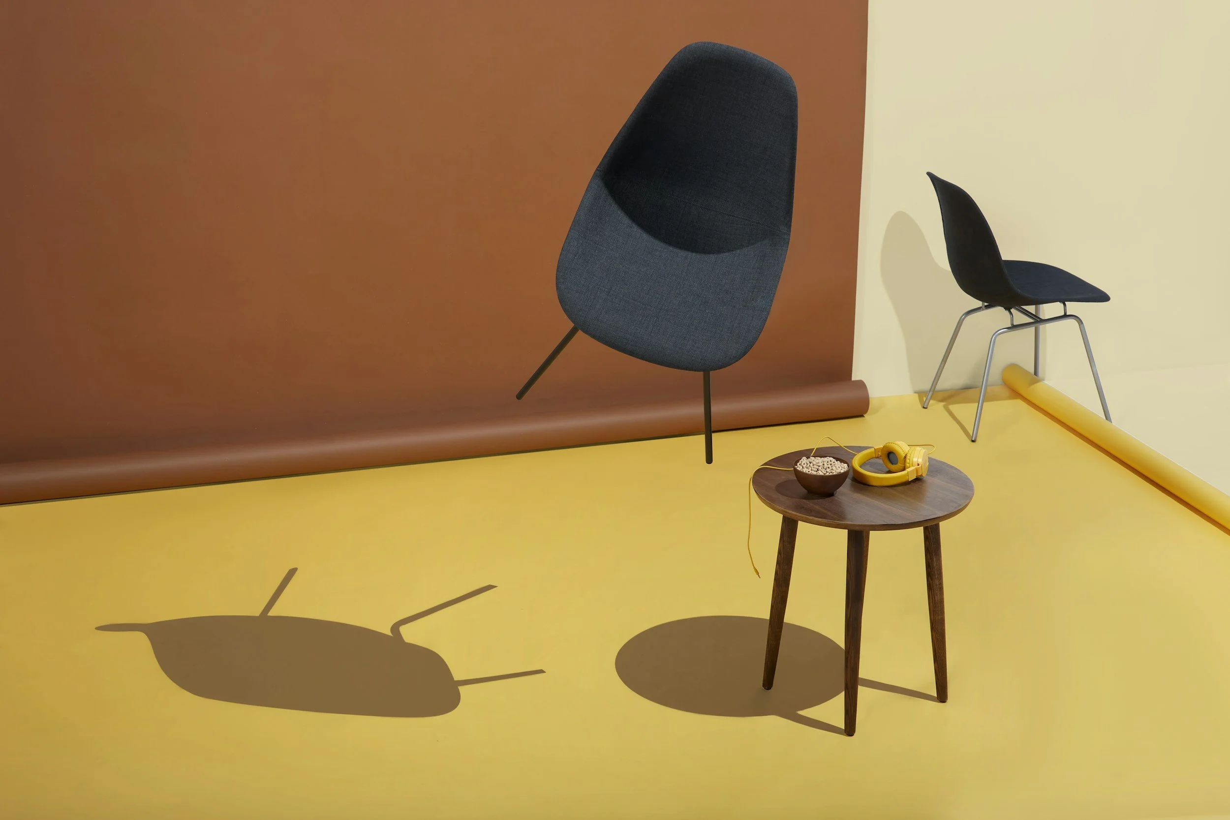 Interior scene with a yellow floor, brown and beige walls, a wooden side table with headphones, cereal, and a bowl, and two modern chairs casting shadows.
