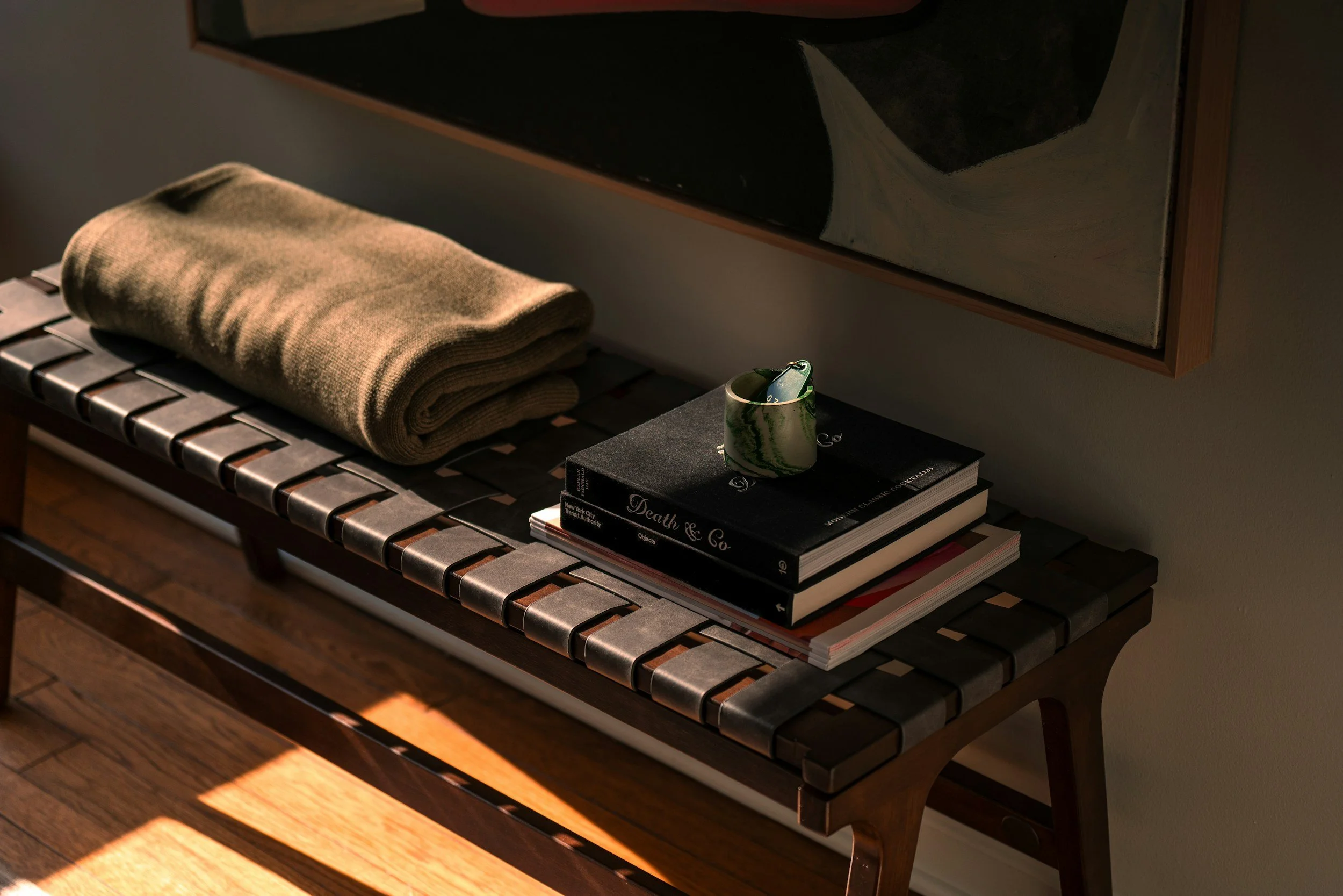 A wooden bench with a woven top, a folded beige blanket, and a stack of books including one titled 'Death & Co.' and a ceramic mug with green marble pattern containing pens or pencils, placed in a dimly lit room.