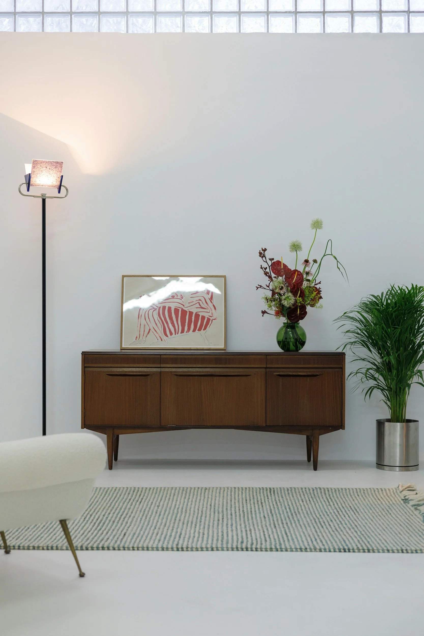 A room with a mid-century modern wooden sideboard, a framed zebra artwork on top, a vase with a floral arrangement, a green leafy plant in a silver pot, a white armchair, a striped rug, and a wall with glass block windows and a tall floor lamp