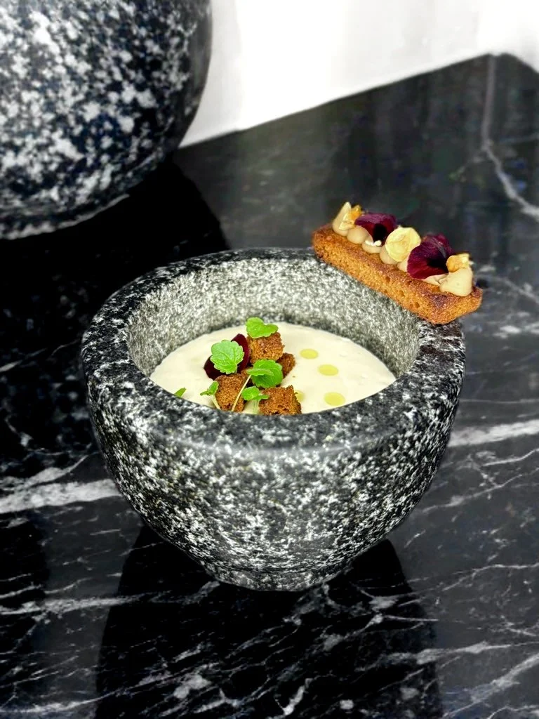 Soupe dans un bol en pierre noire, garnie de morceaux de pain, de petits légumes verts et de gouttes d'huile, accompagnée d'une tranche de gâteau avec des fleurs et des noix sur le dessus, sur une table en marbre noir.