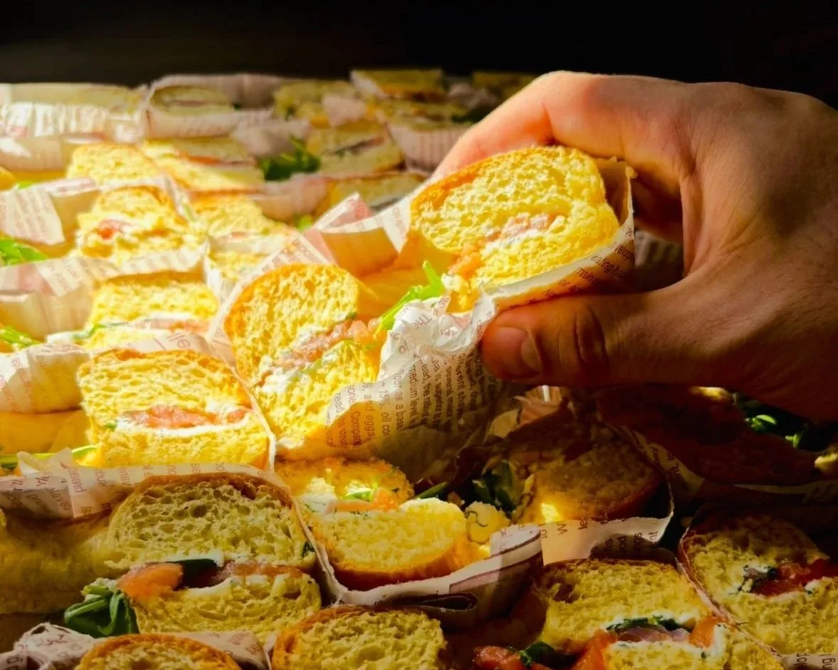 Une main tient un sandwich baigné de lumière, entouré d'autres sandwiches similaires, contenant du pain, des légumes verts, du fromage et de la viande, présentés dans des emballages en papier.