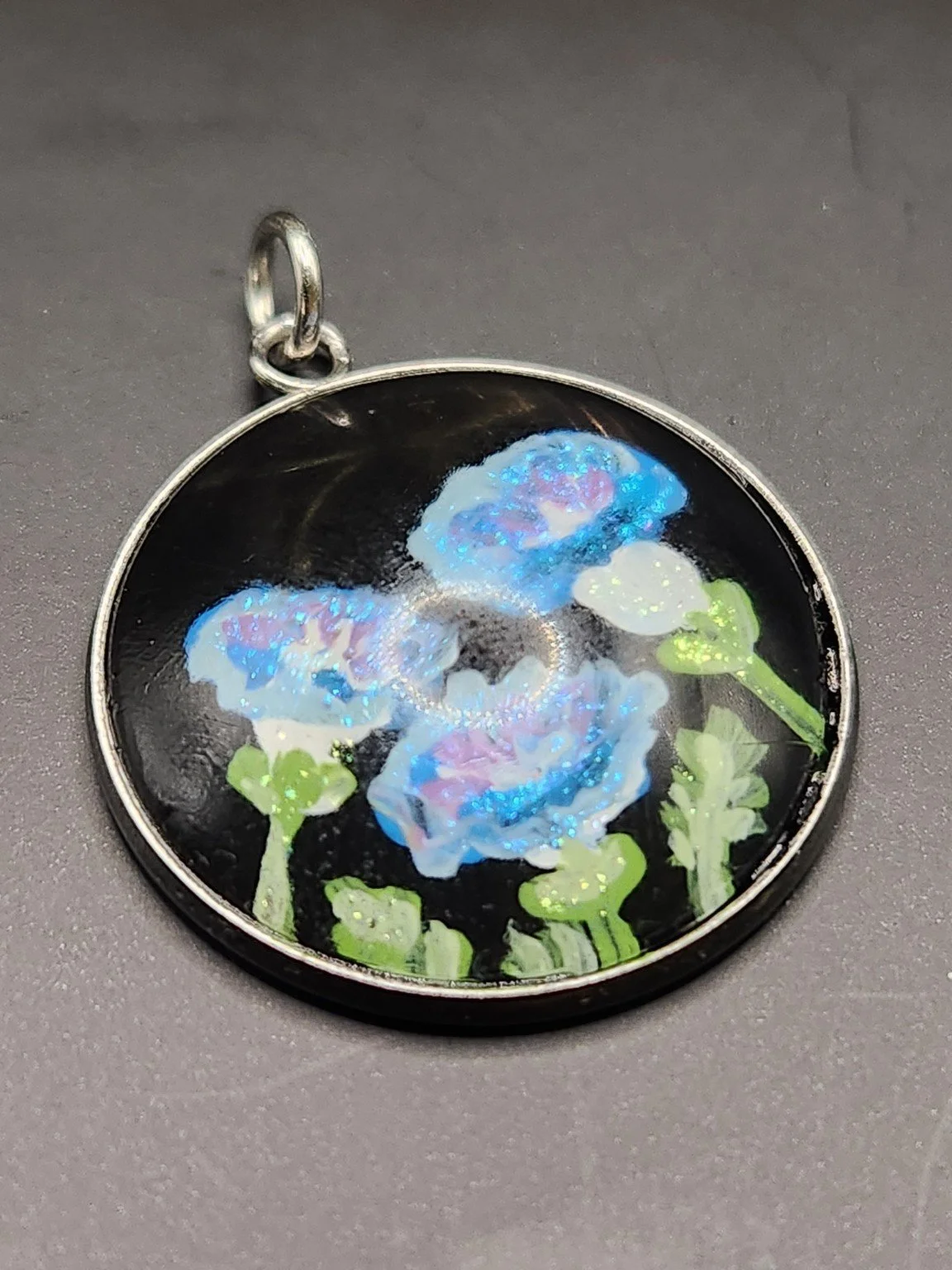 Blue morning glory art pendant with pink centers, green stems, and glitter accents in a silver-tone bezel against a gray background.