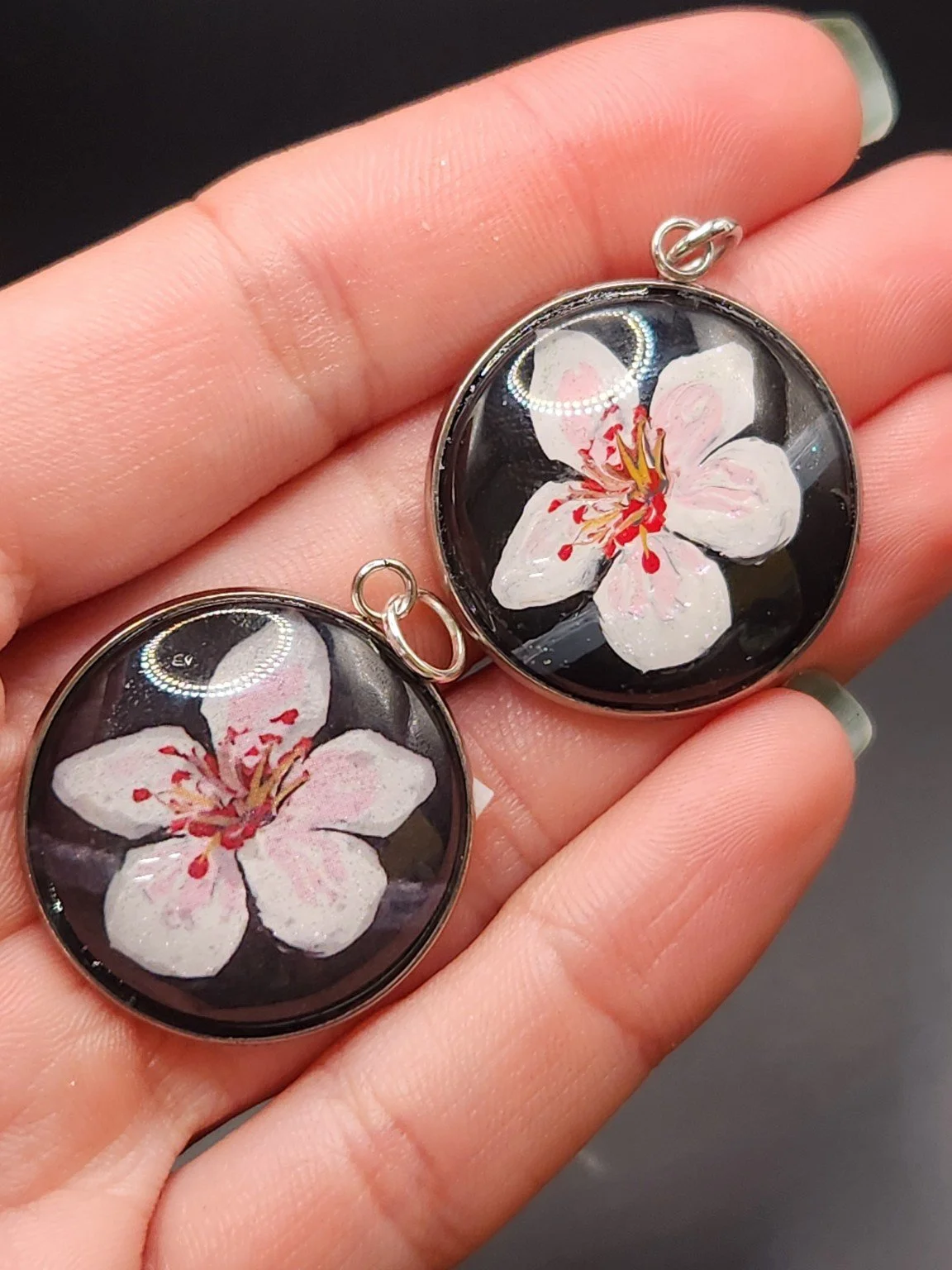 Two cherry blossom pendants in a hand: hand-painted original at the top right and printed reproduction at the bottom left.