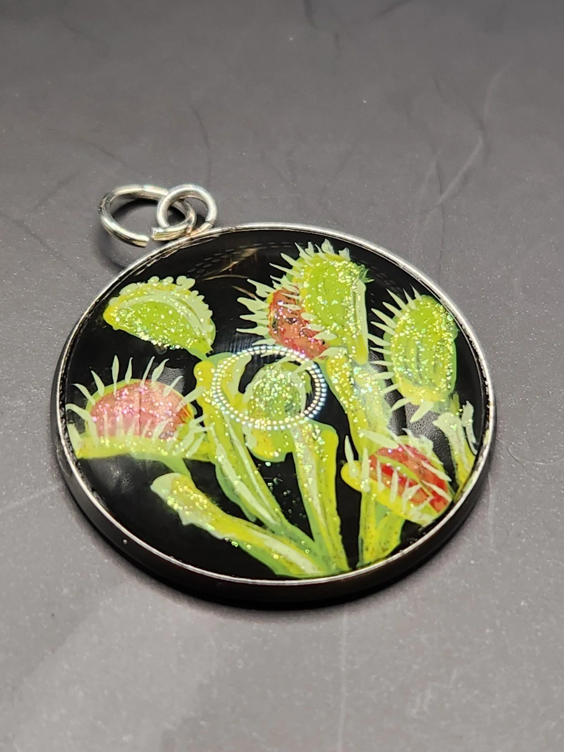 Venus flytrap art pendant with bright green traps, red centers, and shimmering glitter in a silver-tone bezel against a gray background.