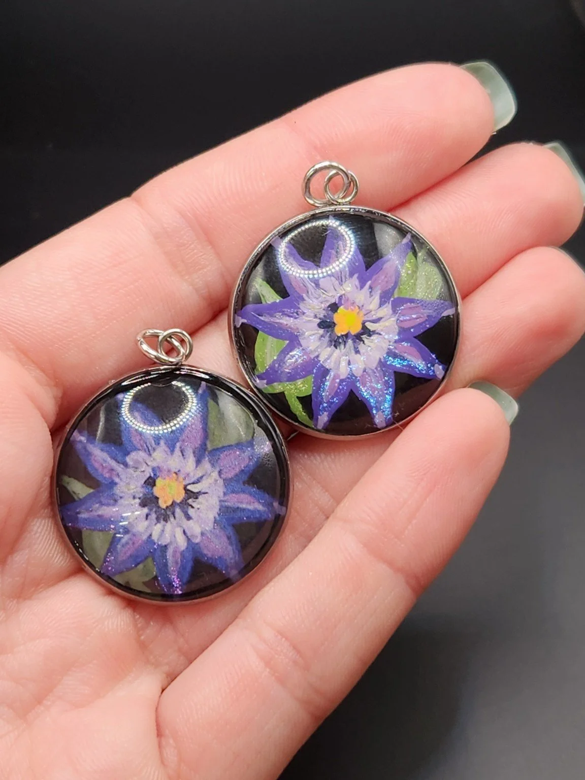 Two clematis flower pendants held in a hand: hand-painted original at the top right and printed reproduction at the bottom left.
