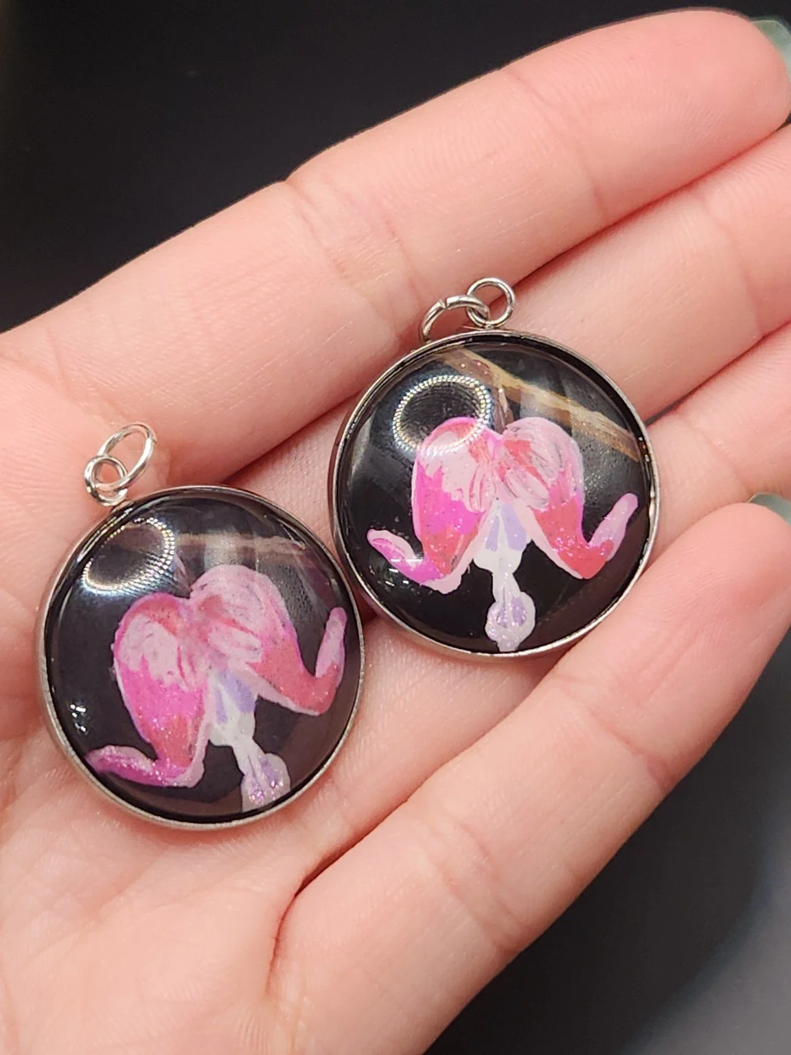 Two bleeding heart flower pendants held in a hand: hand-painted original at the top right and printed reproduction at the bottom left.