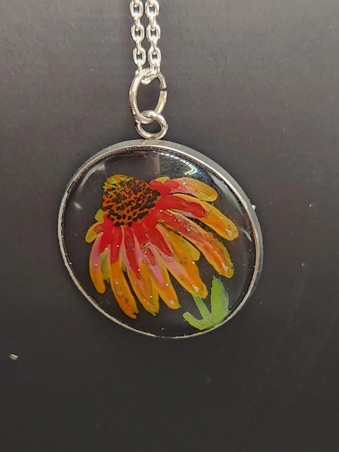 Red and yellow coneflower art pendant shown on a silver-tone chain against a dark background. Chain not included.