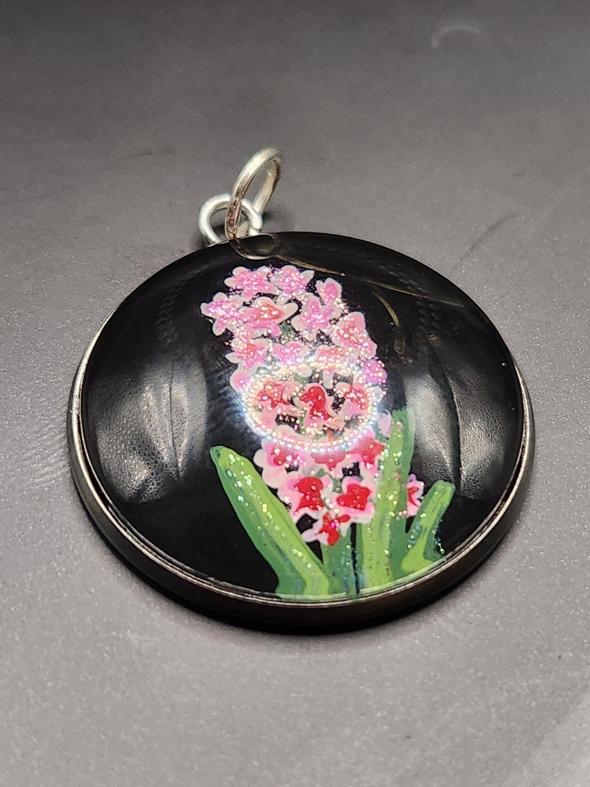Pink hyacinth flower art pendant with shimmering glitter, green leaves, and a silver-tone bezel against a gray background.