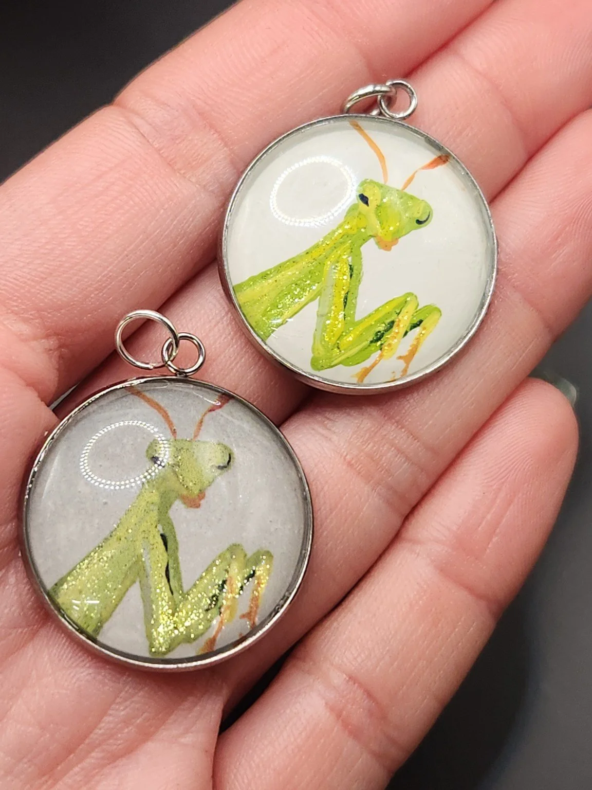 Praying mantis pendant comparison held in a hand: the hand-painted original is at the top right, and the printed reproduction is at the bottom left.