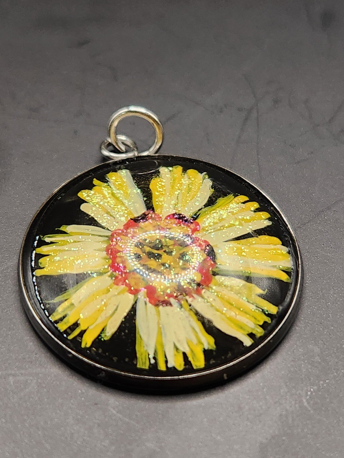Yellow coreopsis flower art pendant with an orange-red center and glitter accents in a silver-tone bezel against a gray background.