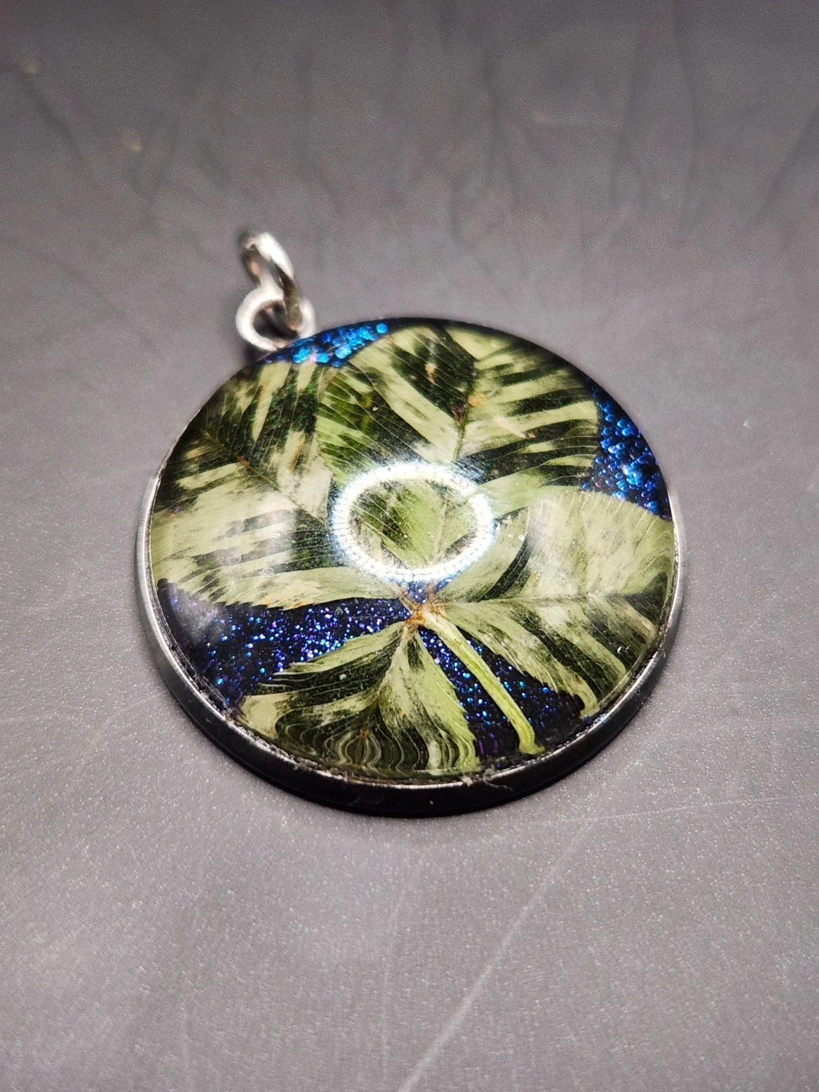 Perfectly Imperfect I004: real four-leaf clover pendant with blue glitter and fern-like translucent leaf patterns, close-up on gray.