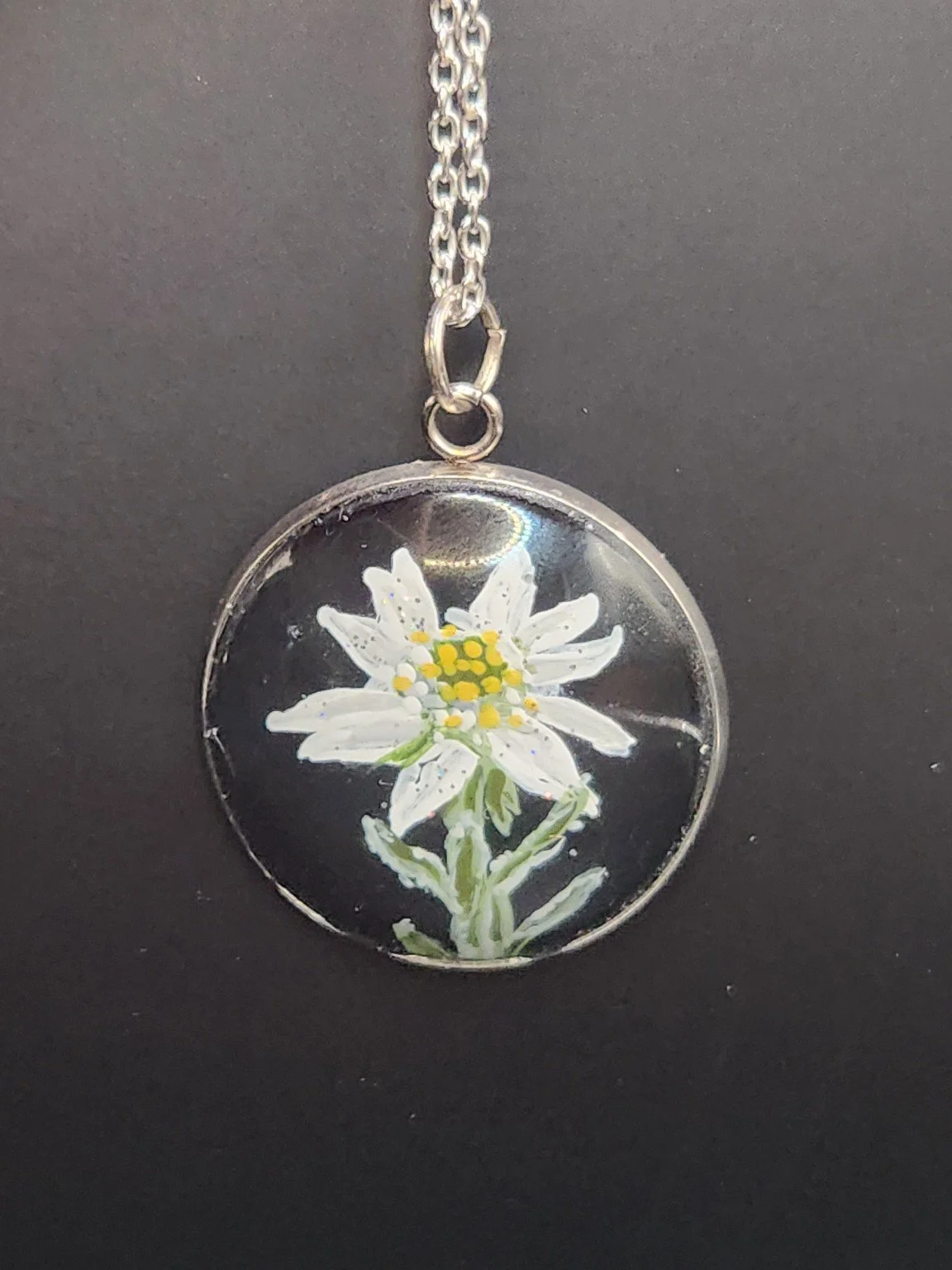 White edelweiss flower art pendant shown on a silver-tone chain against a dark background. Chain not included.